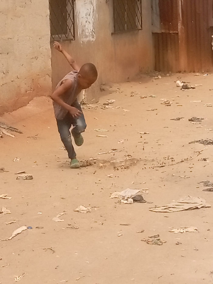 The image presents a daylight scene in Atani, Nigeria, focusing on a young male child in dynamic motion on an unpaved, sandy ground. The boy, with short dark hair, wears a light-colored, sleeveless top, blue jeans, and light green closed-toe shoes. He is captured mid-stride, leaning forward, with his left leg raised and bent, and his right arm extended backward and upward, suggesting running or a similar athletic action. His head is tilted down. The ground is dry, light brown, and heavily scattered with various debris, including white paper scraps, flattened cardboard, and small rocks. In the background, a light-colored building with two windows fitted with dark metal grates is visible to the left. To the right background, a section of a structure made from rusty, corrugated metal sheeting is present.