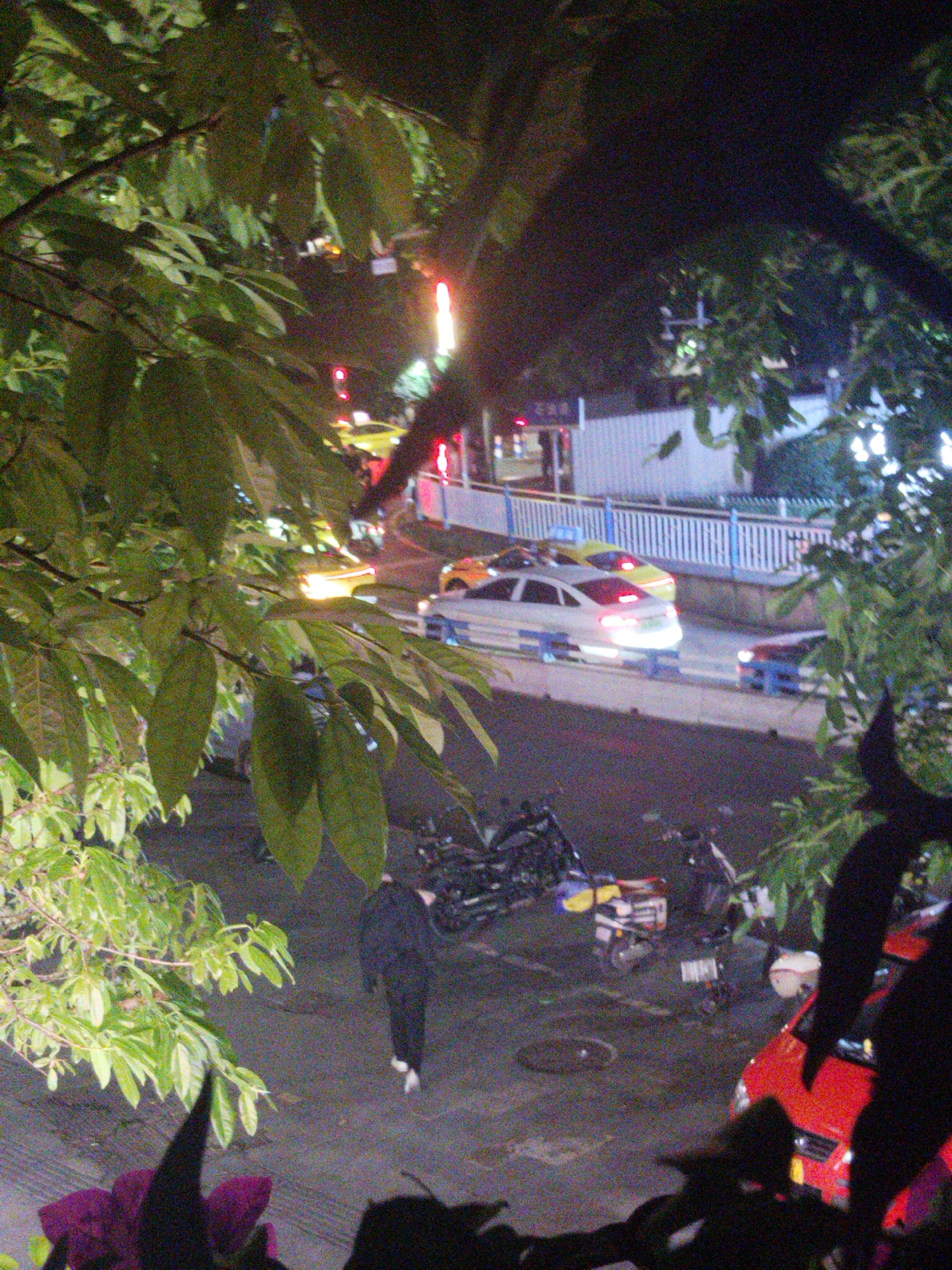 The image, taken at night, shows a street scene in the Yuzhong District, China. A person in dark clothing and light shoes is walking away from the viewer on a pavement littered with parked motorbikes and bicycles. Cars are parked along a road separated by a low white fence. Traffic signals are visible in the background. Leaves from trees frame the image's left and right sides, partially obscuring the view.