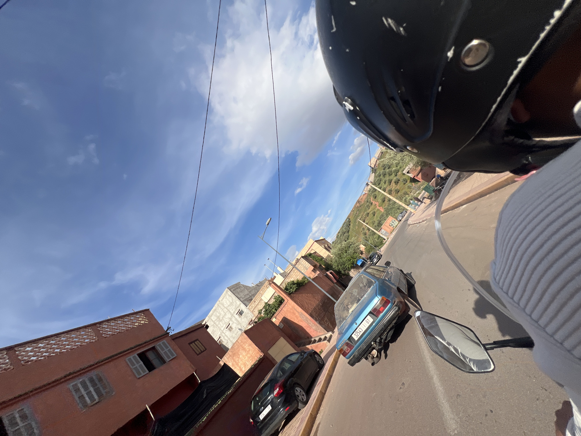 The image, taken from the perspective of a person riding a motorcycle, shows a street scene in Ouaouizert, Morocco. A black helmet is prominent in the foreground, partially obscuring the view. The street curves slightly to the right. Parked along the side of the road are a blue, older model car with a visible license plate number "32543 T 61", and a black hatchback car. Buildings in the background are predominantly reddish-brown and light gray in color. Utility lines are visible against a blue sky with scattered clouds. A hillside with olive trees is also visible in the background. The road surface appears to be asphalt. The motorcycle's rearview mirror is partially visible on the left.