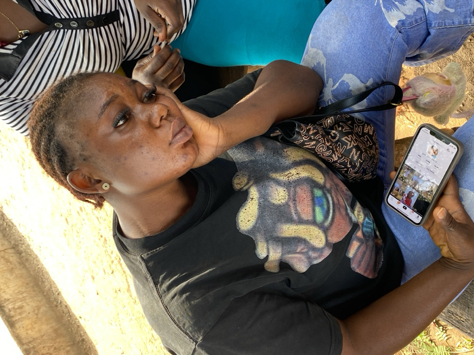 A person rests their chin on their hand while looking at a smartphone. The phone displays a social media profile with several videos or photos. The person wears a black T-shirt with a large graphic on it and sits next to someone in a black and white striped shirt. Light blue jeans are also visible. A black handbag sits on the person's lap. The setting appears to be outdoors in daylight.