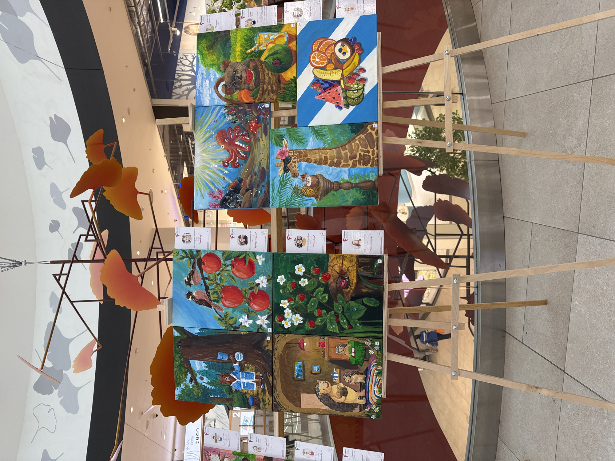 An indoor art exhibition showcases numerous vibrant paintings displayed on simple wooden easels within a modern, multi-level public space, possibly a shopping mall or gallery. The setting features light-colored tiled floors, reflective surfaces, and contemporary architectural design. Large, abstract, orange ginkgo leaf sculptures are suspended from an overhead structure, complementing the display.
The paintings, likely children's artwork, are diverse in subject matter and style. On the top easel, four paintings are visible. The top-left portrays a brown, dog-like creature in a basket filled with watermelon and other fruits in a green landscape. The top-right features an arrangement of various fruits like oranges, bananas, berries, kiwi, and watermelon against a blue and white striped background. Below these, a red octopus is depicted in a bright underwater scene, and next to it, a giraffe wearing sunglasses and a flower poses beside a pineapple.
On the lower easel, another set of four paintings is displayed. The top-left shows two birds on branches adorned with red apples and white blossoms. Beside it, a lively scene of strawberries with white flowers, featuring a ladybug. The bottom-left painting illustrates a person in a blue robe outside a treehouse in a forest. Finally, the bottom-right presents anthropomorphic hedgehog characters inside a cozy, burrow-like home.
Each painting is accompanied by a small card, likely identifying the artist, often with a small portrait and Cyrillic text, indicating the exhibition is in a Russian-speaking region, consistent with the provided context of Perm, Russia. The scene is brightly lit by indoor lighting, and the overall impression is one of creative, youthful artistry.