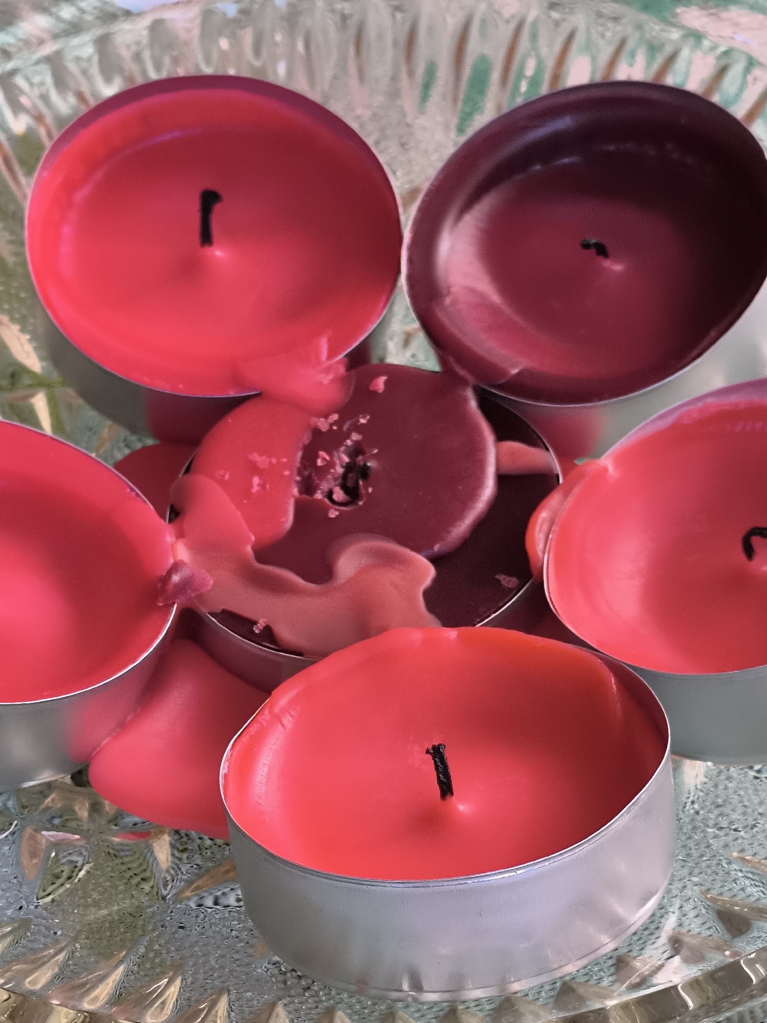 The image shows a close-up, top-down view of several melted red and dark red tea light candles, some still in their silver metal containers, arranged on a decorative, textured glass surface. The candles are in various states of melt, with wax pooling and dripping. One candle has a partially burned wick visible. The background is out of focus, showing the texture of the glass and some green and light-colored elements, possibly foliage or fabric. There are no people or animals in the image. The scene is a still life of melted candles. The location of Dercekliai, Lithuania is not depicted or suggested by any elements in the image.
