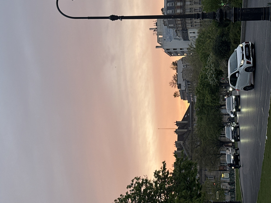 The image depicts an urban street scene in Paris, France, at twilight or dusk. The sky transitions from a light blue at the zenith to vibrant orange and pink hues near the horizon, indicative of sunset. A dark, ornate lamppost stands vertically on the far right, partially cropped.
In the foreground and mid-ground, a multi-lane asphalt road stretches towards the background, with a grassy median strip separating traffic directions. Five vehicles are visible on the road, all with headlights illuminated. In the rightmost lane, a white Kia Picanto (bearing French license plate "FM-090-DN") drives away from the viewer. Behind it, a dark sedan, possibly a Mercedes-Benz, also proceeds with headlights on. Further back in the same lane, another dark car with active headlights is visible. In the leftmost lane, a dark SUV or car with headlights on moves away from the viewer. A lone individual on a bicycle, wearing dark attire, rides behind this SUV in the leftmost lane. On the far left, a distant group of indistinct figures can be seen on a sidewalk, near a rack of public bicycles (likely a Vélib' station).
The background is dominated by typical Parisian architecture. On the left, several multi-story buildings feature classic Haussmannian design, characterized by grey slate mansard roofs, numerous chimneys, and dormer windows. One prominent building in the center-left background displays intricate architectural details and a tall, slender spire or antenna on its roof. Further to the right, a taller, more contemporary white building with visible graffiti on its side stands alongside other traditional structures. Several rooftops on various buildings support small clusters of green vegetation. Numerous deciduous trees with full green foliage line the street and are scattered throughout the scene, particularly on the left side and within the median.