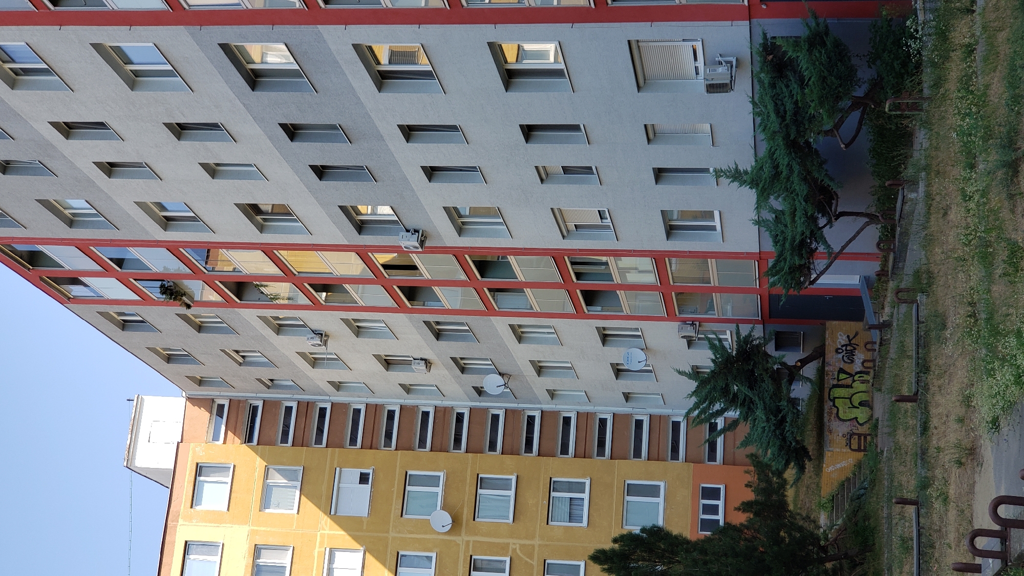 The image shows a residential building in Bratislava, Slovakia. The building is a tall, multi-story structure with a gray facade and red accents around the balconies. To the left of the gray building is a section of a building with a yellow facade. The ground level appears to be a courtyard with patchy grass and some trees. There is graffiti on a low wall near the entrance to the building. The sky is clear and blue.