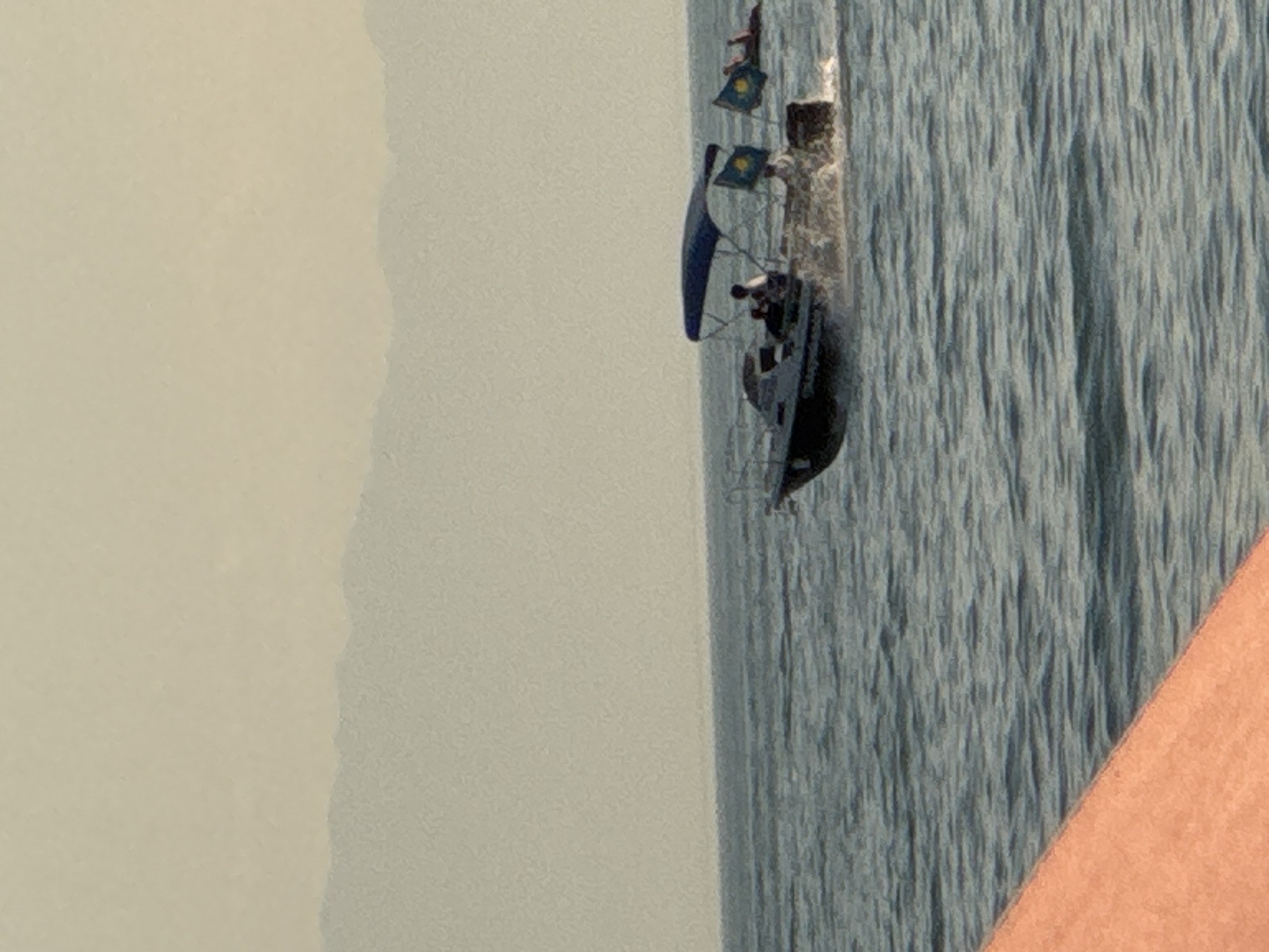 A small motorboat with a blue canopy is navigating on a body of water, leaving a wake. Two yellow flags with blue symbols are attached to the back of the boat. Three individuals are visible on the boat, with one appearing to steer. In the background, a distant, hazy mountain range is visible against a pale sky. To the right of the motorboat, a person is lying on an inflatable tube in the water. A portion of an orange surface, possibly a dock or part of another vessel, is in the foreground, angled from the bottom left. The scene appears to be a recreational setting on the water.