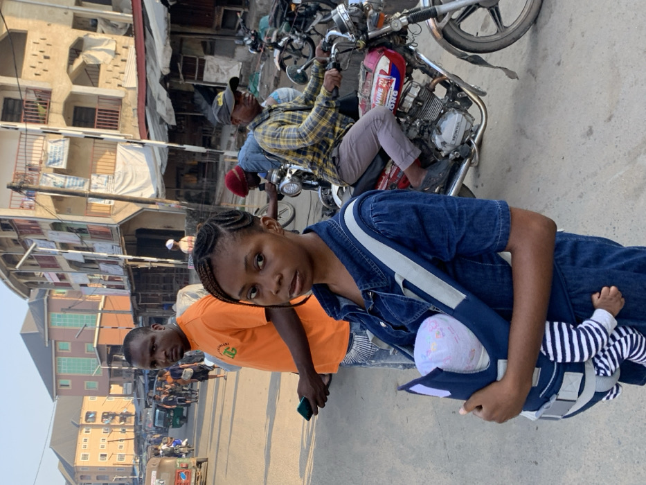 A daylight street scene depicts a bustling urban environment in Aba, Nigeria. In the foreground, a young Black female, with braided hair, wears a denim jumpsuit and faces the camera directly, holding an infant in a navy blue baby carrier strapped to her chest. The infant wears a light cap and striped sleepsuit. Immediately behind her, a Black male in an orange t-shirt and light-colored jeans stands, holding a dark smartphone in his left hand, also looking towards the camera. To the right, a Black male wearing a plaid shirt and dark baseball cap sits astride a red and silver Honda motorcycle, looking forward.
The background features a paved street with other pedestrians, motorcycles, and a green tricycle (Keke Napep) visible further down. On the right, a multi-story building with light-colored walls and balconies is visible, with multiple banners or fabric pieces hanging from it, and what appear to be ground-level market stalls. Further left in the background, additional multi-story buildings with light and orange facades are present. Utility poles and overhead wiring are visible throughout the scene. Shadows cast on the ground suggest a specific time of day with natural sunlight.