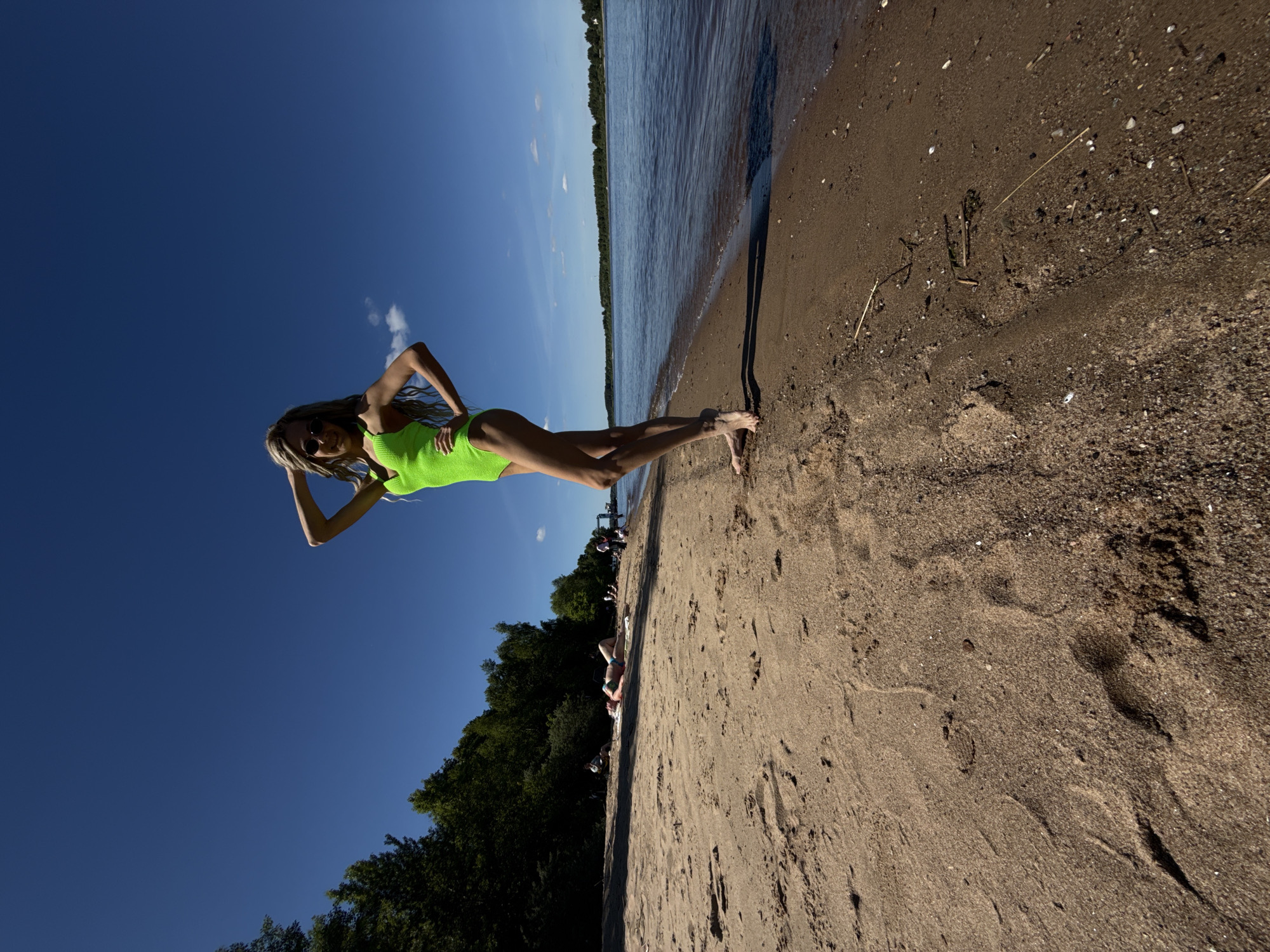 The image depicts a sunny day at a sandy beach in Rusanovo, Russia, adjacent to a calm body of water, likely a river or large lake. A prominent young woman stands barefoot in the foreground on the damp sand at the water's edge, facing the viewer. She is wearing a bright neon green one-piece swimsuit and sunglasses, with long, light-colored hair, part of which is styled into dreadlocks. Her left arm is raised with her hand behind her head, and her right hand is placed on her hip, indicating a posed stance. Her body casts a long shadow on the sand behind her.
In the mid-ground, further down the sandy beach to the left, several other individuals are present. One person is distinctly visible lying supine on a light-colored towel or mat on the sand, wearing a bikini. Other figures are scattered along the treeline and closer to the water. The background on the left is dominated by a dense line of dark green deciduous trees. To the right, the wide expanse of water extends to a distant forested shoreline under a clear, deep blue sky with minimal white clouds. The sandy beach shows various textures, footprints, and scattered small natural debris like sticks.