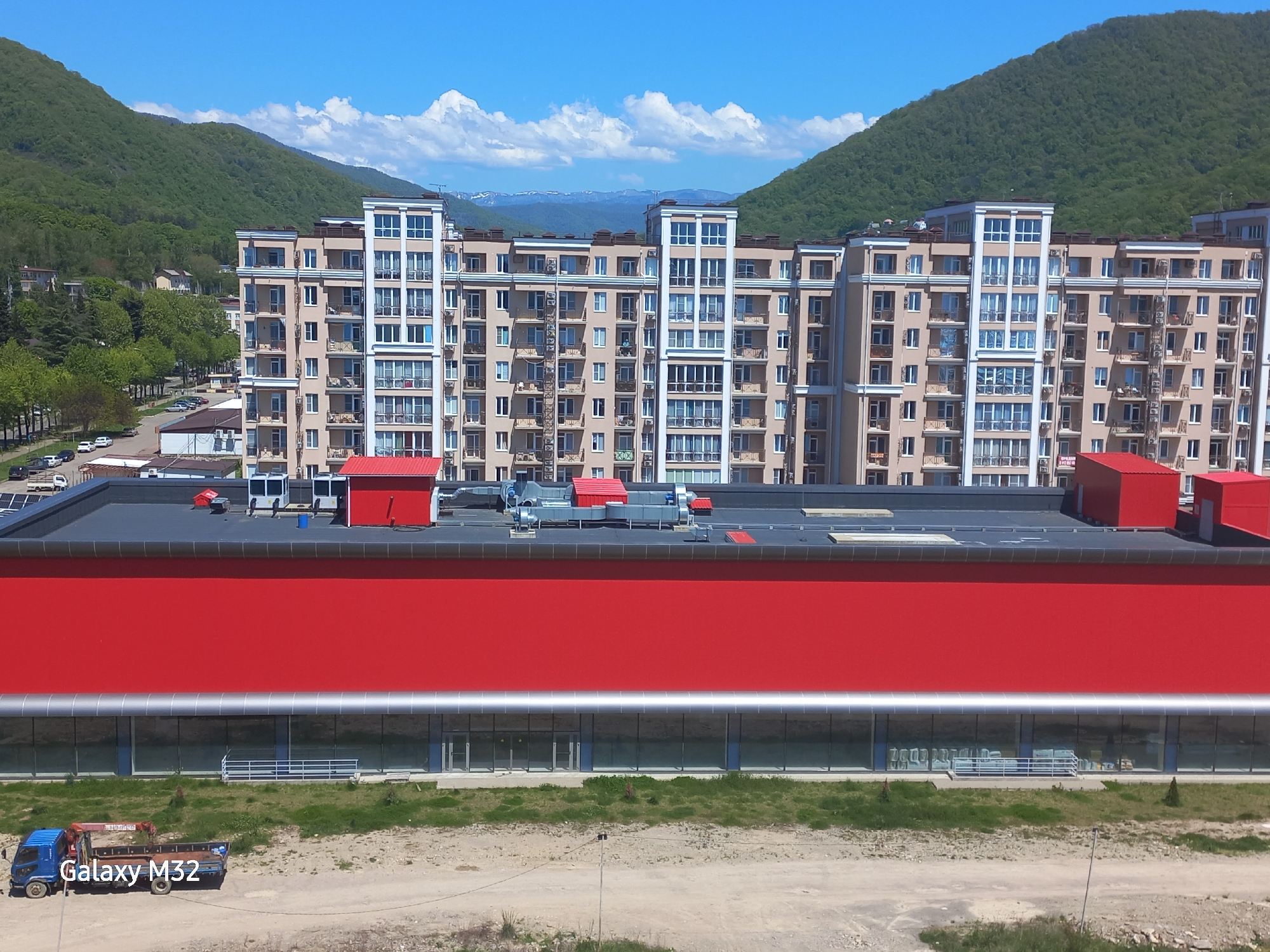 The image depicts a landscape in Sochi, Russia, featuring a red building with glass windows at its base. The red building has a flat roof with several red structures and ventilation equipment. In front of the red building, there is a blue truck parked on a dirt and grassy patch. Behind the red building stands a large, multi-story building with a beige facade, balconies, and white window frames. In the background, mountains rise up, capped with clouds and snow in the distance. The sky is clear and blue. A street with parked cars and trees lines the left side of the image.