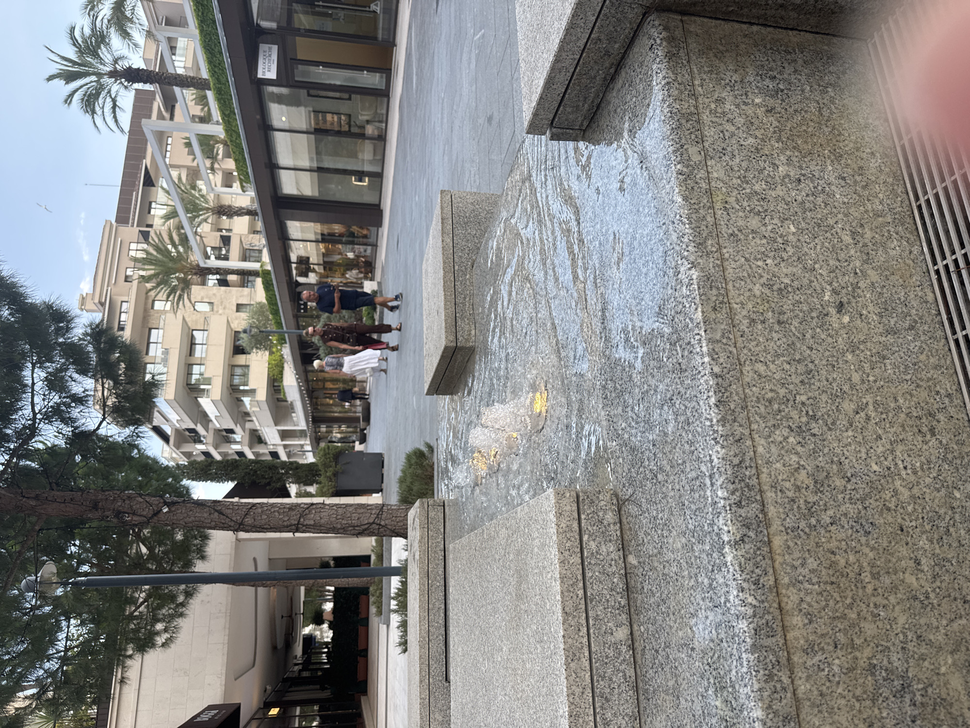 The image depicts an outdoor scene with a modern architectural setting. In the foreground, a tiered water feature made of granite is visible, with water cascading over its surfaces. The stone has a speckled, grey and black pattern. A metal grate is also present on the right side of the foreground.
In the midground, several people are walking on a paved walkway. One person, a man in a dark blue shirt and shorts, is facing left. Two other individuals are further down the path. The walkway is bordered by buildings with large glass windows and modern facades, suggesting a commercial or residential area. Palm trees are interspersed with the buildings, indicating a warm climate.
The background features multi-story buildings with balconies and light-colored exteriors. The sky is visible above, appearing overcast or early evening. The scene appears to be in a developed urban area, possibly a plaza or promenade.
There is text visible on a building in the upper left portion of the image, which appears to read "BIOLOGIQUE RECHERCHE".
The overall impression is one of a clean, contemporary urban environment, possibly in Tivat, Montenegro, with people enjoying the public space. The time of day is likely late afternoon or early evening.