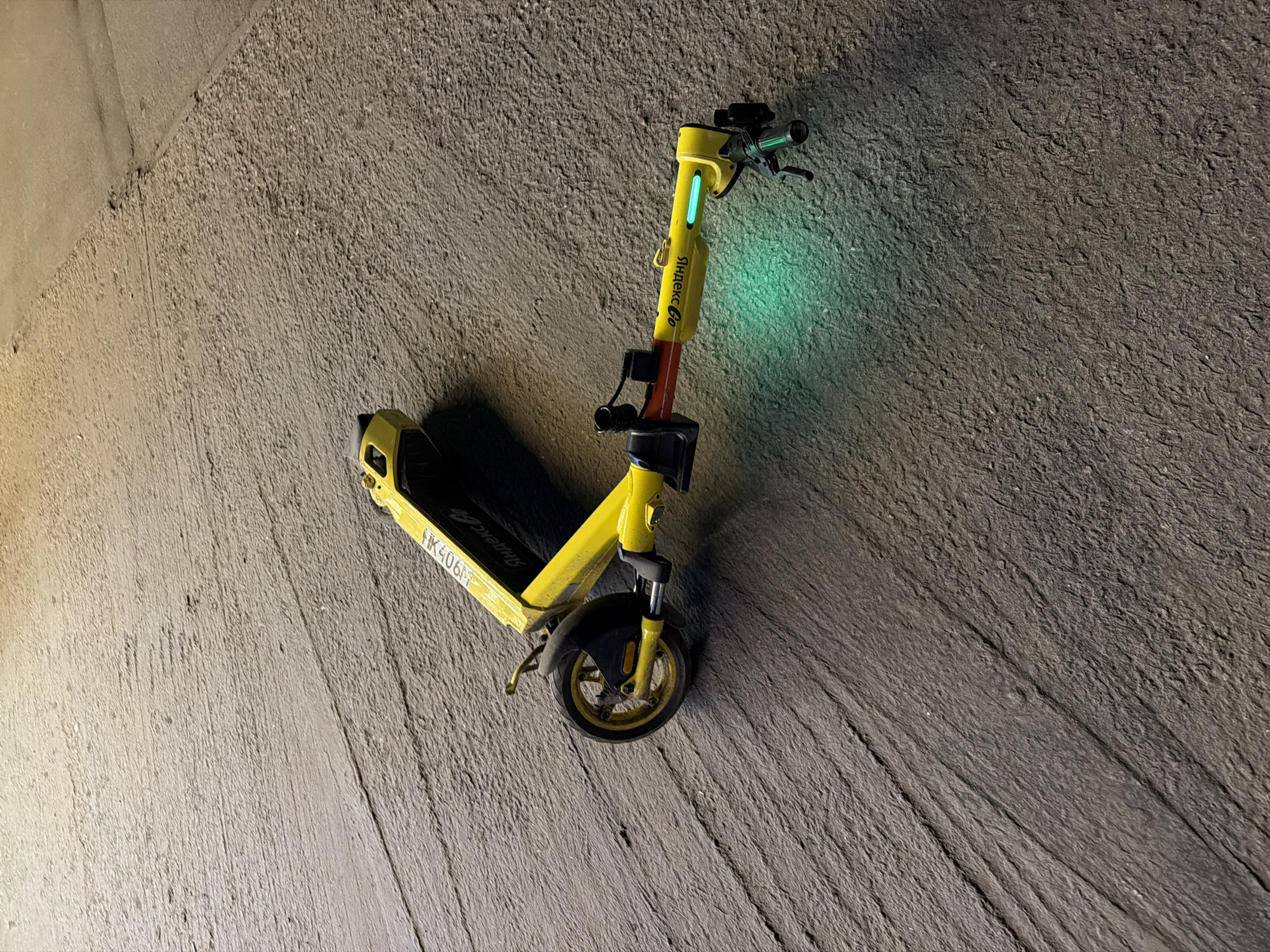 A bright yellow electric scooter is the central subject of this image, positioned diagonally against a textured, dark gray concrete surface. The scooter features a prominent green light emanating from its stem, casting an ethereal glow onto the adjacent concrete.
The scooter is primarily yellow, with black details on its wheels and handlebar grips. Text on the scooter's stem and footboard clearly displays "Яндекс Go" (Yandex Go), identifying it as part of that service. A serial number or identifier, "HK406M," is also visible on the footboard near the front. The scooter has a front wheel with a black fender and a kickstand.
The background is a rough, uneven concrete surface, characterized by a distinct grooved or raked texture that runs diagonally across the frame, complementing the scooter's angle. The scene is dimly lit, suggesting either nighttime or an enclosed, dark environment like an underpass or garage. No activity is taking place; the scooter appears stationary. The location is Khimki, Russia.