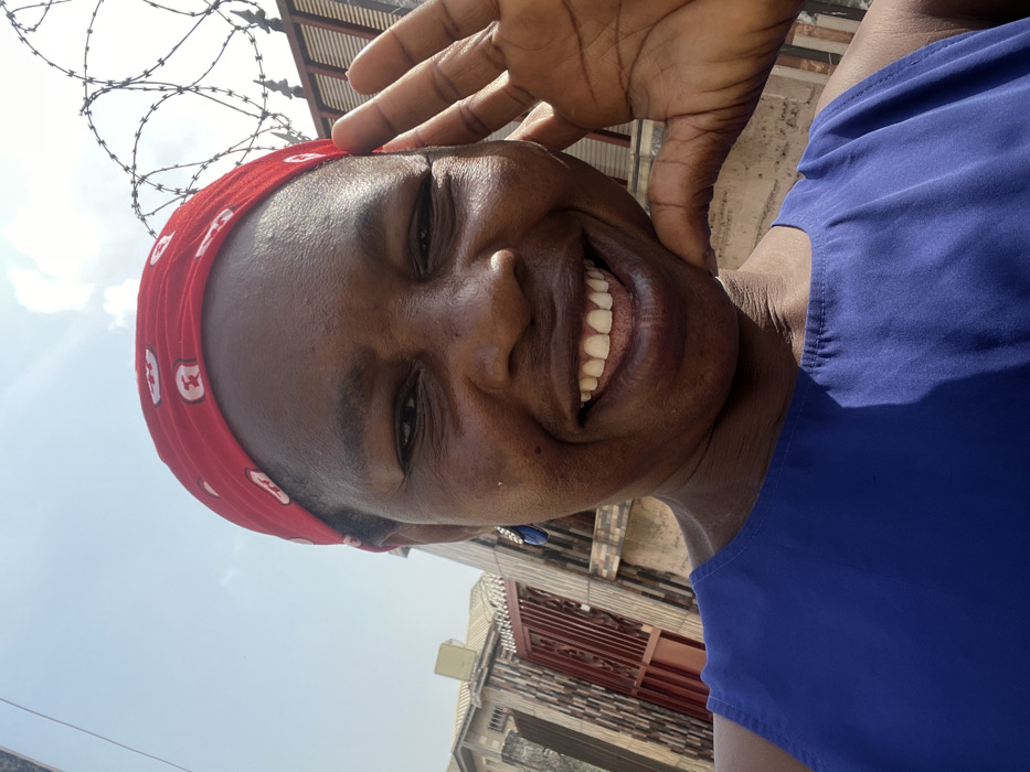 The image is an outdoor, close-up selfie of an adult female, depicting a scene in Iranla, Nigeria. She has dark skin and is smiling broadly, revealing her teeth. Her head is covered by a red fabric headwrap adorned with white patterns. She is wearing a royal blue sleeveless top, and a blue drop earring with a metallic detail is visible on her left ear. Her right hand is partially shown, positioned near her jawline with fingers slightly spread. The background features a clear blue sky with sparse white clouds. Visible urban elements include a light-colored building, a brown metal gate, and a textured wall on the left side. On the right, a darker fence or gate is topped with multiple coils of razor wire.