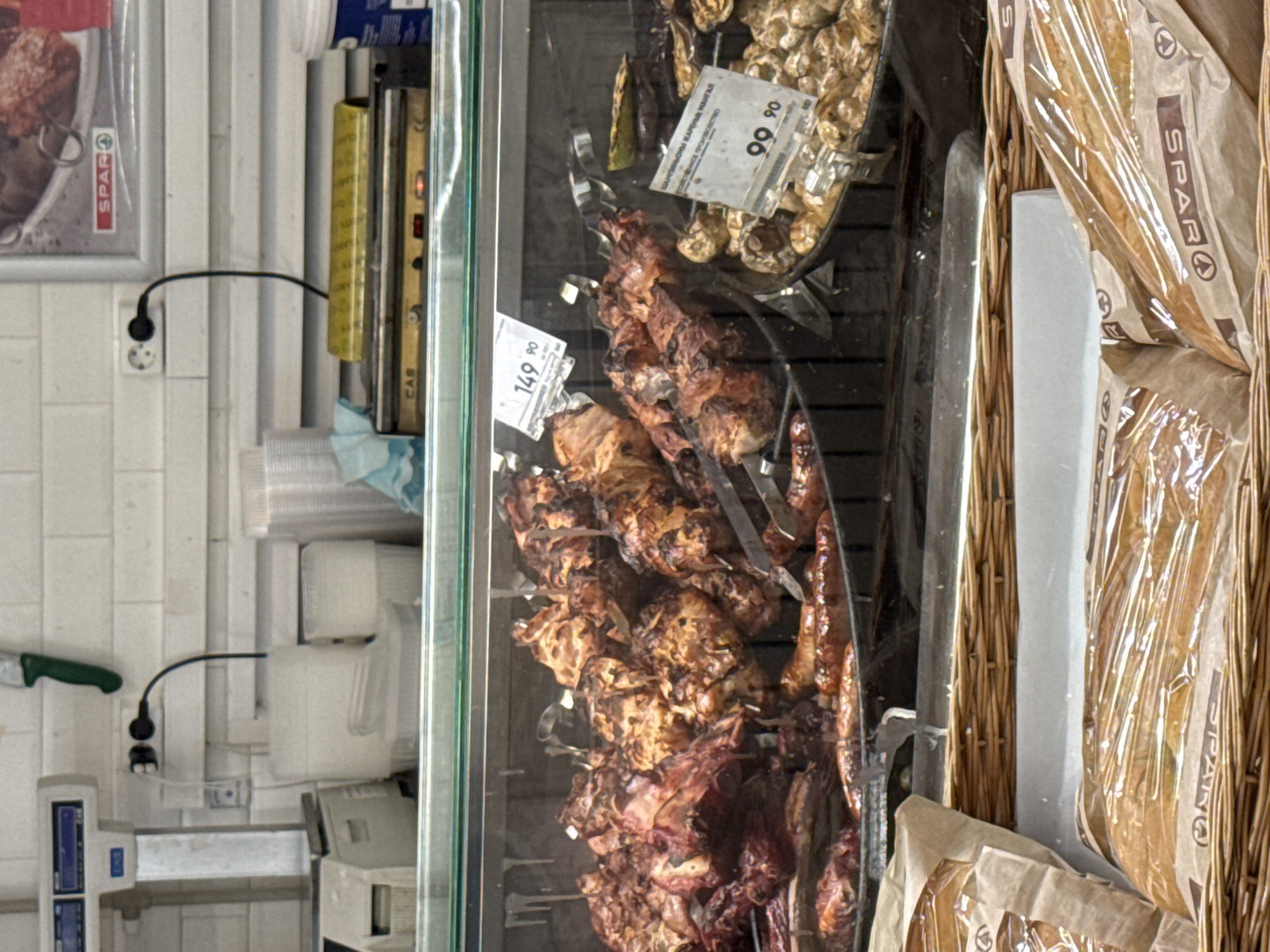The image shows a close-up view of a display case filled with grilled meat skewers and bread. The meat skewers, some appearing to be chicken and others pork, are seasoned and cooked to a golden brown. They are arranged vertically on metal rods, suggesting they are part of a rotisserie or a similar cooking setup. To the right of the meat, several loaves of bread are visible, wrapped in clear plastic packaging with brown paper accents. These loaves are housed in a wicker basket, adding a rustic touch to the display.
In the background, a section of the store's wall is visible, tiled in white. A black electrical cord is connected to a white wall socket, indicating the presence of power for the display. There are also stacks of white plastic containers and a green-handled knife on a shelf. A price tag is visible on one of the meat skewers, with the text "149 90" printed on it, likely indicating the price in rubles. Another tag above the bread displays "99 90". The overall setting appears to be a food retail environment, possibly a supermarket or a deli counter, given the presence of food items and display fixtures. The lighting is bright, suggesting it is daytime or the store is well-lit. There are no people visible in the image, focusing solely on the products. The brand "SPAR" is visible on some of the packaging, identifying the store or brand. The image seems to be taken from a slightly elevated angle, looking down into the display.