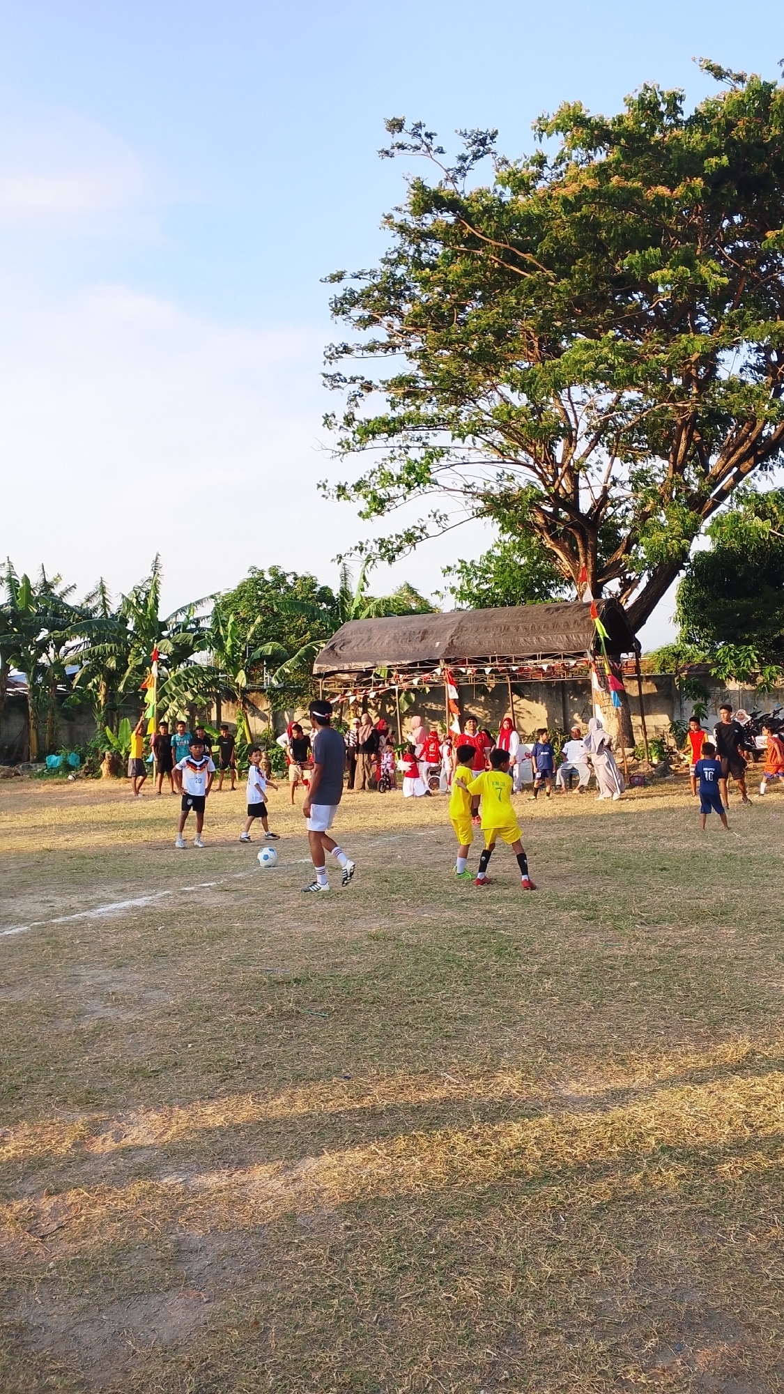 In the city of Unknown, Indonesia, an outdoor football match is underway on a dry, grass-covered field. The sun casts long shadows, indicating it is late afternoon or early evening. A crowd of spectators has gathered to watch the game, some seated under a makeshift canopy decorated with Indonesian flags. Children and adults are participating in the football match, some wearing team jerseys and shorts, while others are in casual attire. The background features lush greenery from banana trees and a large, mature tree, suggesting a tropical or subtropical environment. The atmosphere appears festive and communal, with families and friends enjoying a sporting event.