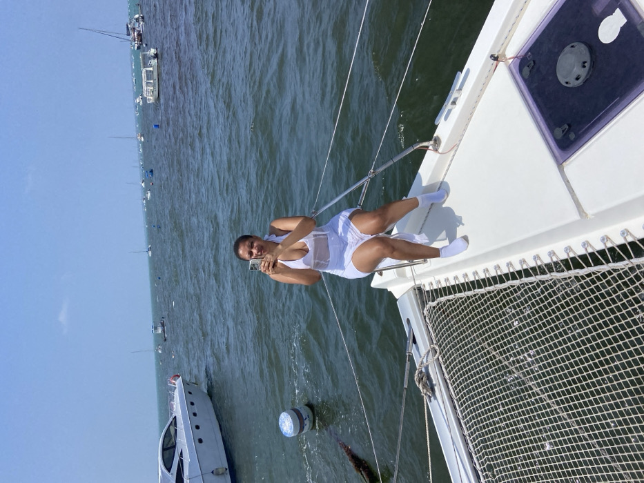 The image shows a woman in a white dress sitting on a white catamaran boat, holding a cell phone and taking a picture, with her legs outstretched in front of her. The boat is docked in the harbor of Dar es Salaam, Tanzania. The location is determined by the coordinates, which are -6.752 latitude and 39.272 longitude. In the background, there are several other boats, including a larger white boat that appears to be a yacht, as well as a blue buoy. The water is calm and blue. The sky is clear and blue. The scene is bright and sunny, and the woman appears to be enjoying the beautiful day.