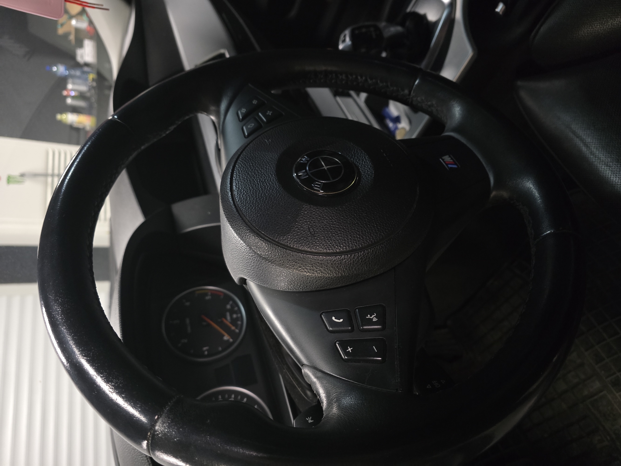 The image shows the interior of a car, specifically the steering wheel and dashboard area. The steering wheel is black leather and shows signs of wear. There are buttons on the steering wheel, including volume controls, a phone button, and a voice command button. A BMW logo is visible in the center of the steering wheel. The dashboard includes a tachometer and speedometer. The overall lighting suggests it is either nighttime or the car is in a dimly lit area. The image does not provide any information about the location in the city of Ruse, Bulgaria. There are no people visible in the image, and no specific actions or interactions are depicted. The environment is that of a car interior.