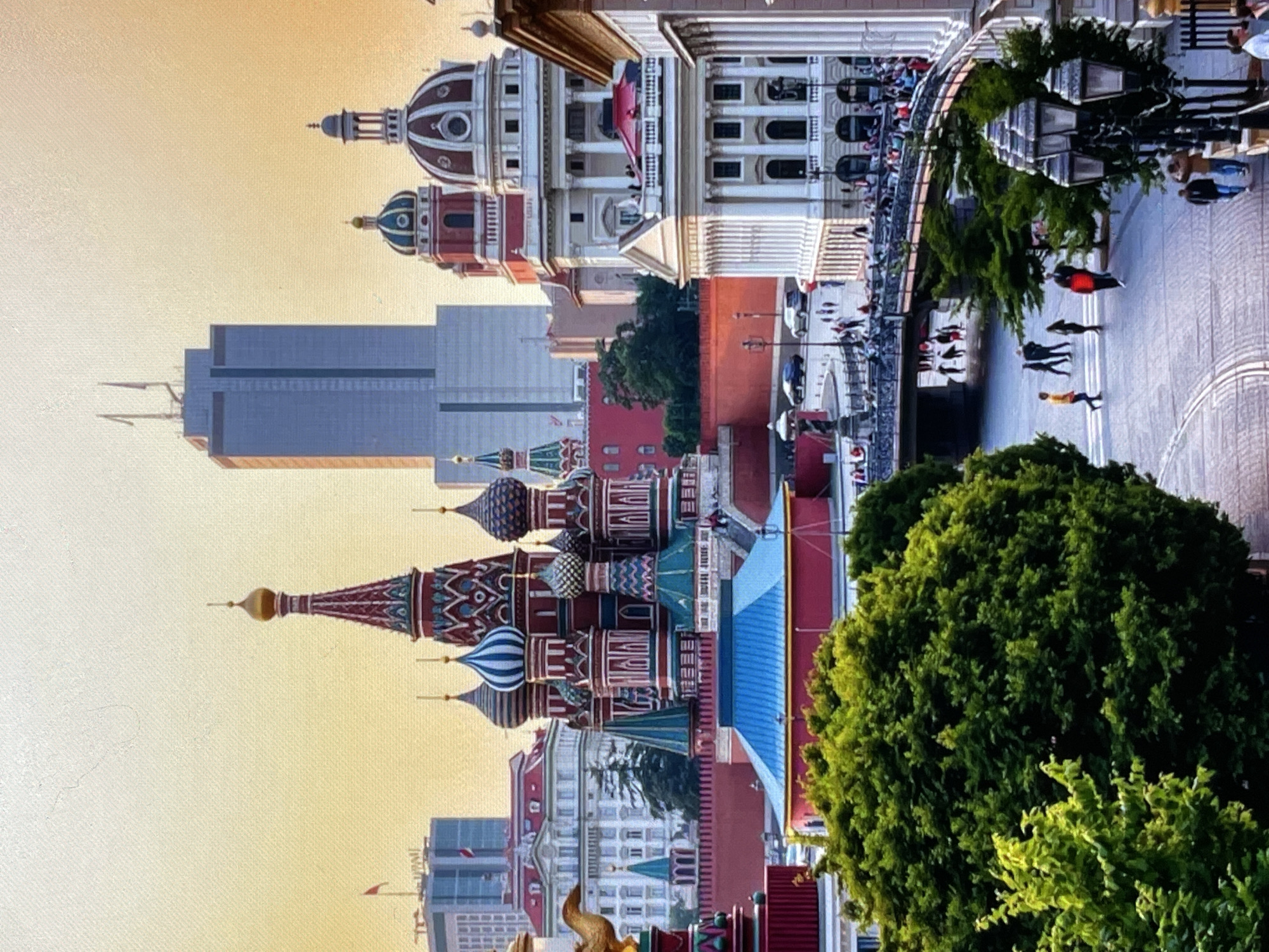 The image depicts a scenic cityscape with prominent architecture and a clear sky transitioning to a warm hue, suggesting either sunrise or sunset. The foreground is dominated by lush green trees, with a large, dense tree on the left and a smaller, brighter green tree in the lower left corner.
In the mid-ground, a vibrant, red-walled structure is visible, featuring a distinctive blue-roofed building. To the right of this, an ornate, multi-domed cathedral, characteristic of Russian architecture, stands tall with its colorful onion domes. Further to the right, several other buildings are visible, including a grand white neoclassical building with a dome and columns, and another building with a red roof. A bridge with railings spans over a lower area, populated by many pedestrians. Cars are also present on a road to the left of the bridge.
In the background, a modern, tall, rectangular skyscraper with many windows dominates the skyline. To the left of this skyscraper, another taller building with a flagpole is visible. In the far distance, more buildings and structures can be seen.
The scene is populated with numerous individuals. Several people are walking on the bridge and the street. In the mid-ground, a few figures are seen near the buildings. In the foreground, there are a few individuals walking on the paved area, and a cluster of people near the lampposts on the right. The overall atmosphere is that of a bustling urban environment with a mix of historical and modern architecture, set against a picturesque sky. The location appears to be Red Square in Moscow, Russia, given the iconic St. Basil's Cathedral and the Kremlin walls, not Pushkino, Russia.