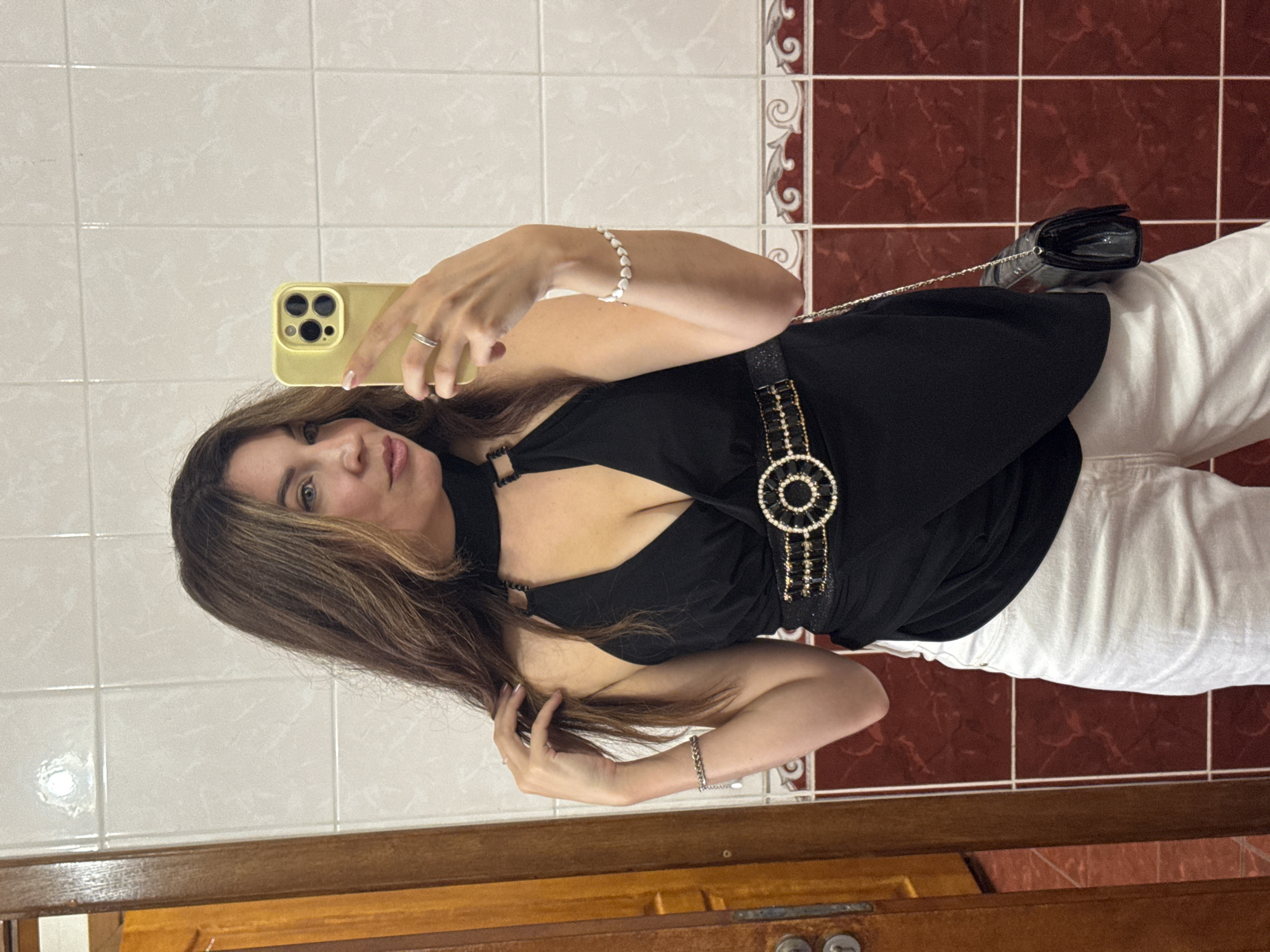 A woman with long, brown hair takes a selfie in a bathroom. She is wearing a black, halter-neck top with a plunging neckline and a sparkly belt. She has on white pants and is holding a yellow phone in her right hand. A black clutch purse hangs from a chain on her left side. The bathroom has white and red tiled walls. The woman is likely in the city of Kryvyi Rih, Ukraine, though there is no explicit indication of this in the image.