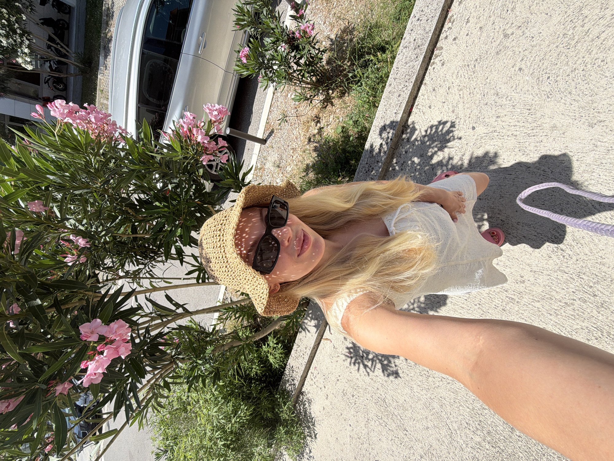 A blonde woman wearing a straw hat, sunglasses, and a white dress poses for a selfie in Budva, Montenegro. She is kneeling on a textured concrete surface, with her left arm extended towards the camera. Behind her, a tree with pink flowers is in bloom. A silver car and parked motorcycles are visible in the background, along with buildings and a street. The scene is illuminated by sunlight, casting shadows on the ground.