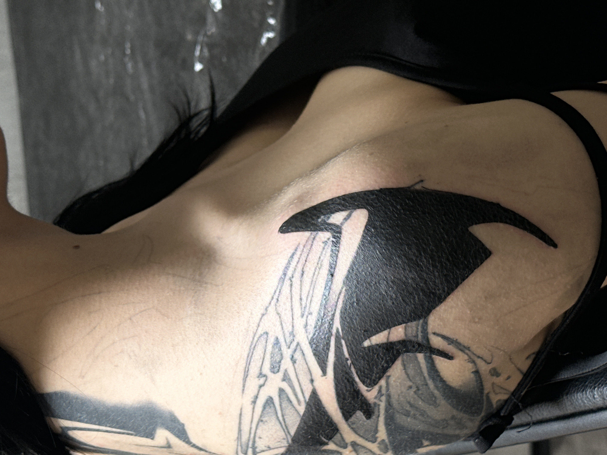 A person is lying down, likely on their back, with the left side of their chest and upper shoulder visible. The person has a black tattoo covering a large portion of their chest and shoulder area. The tattoo appears to be composed of bold, dark shapes and thinner, more intricate line work. The person is wearing a black garment that covers part of the neck and shoulder, with a thin black strap visible near the right shoulder. The background is a dark, textured surface, possibly the seat or back of a chair or examination table. The location in Moscow, Russia is not evident from the image.