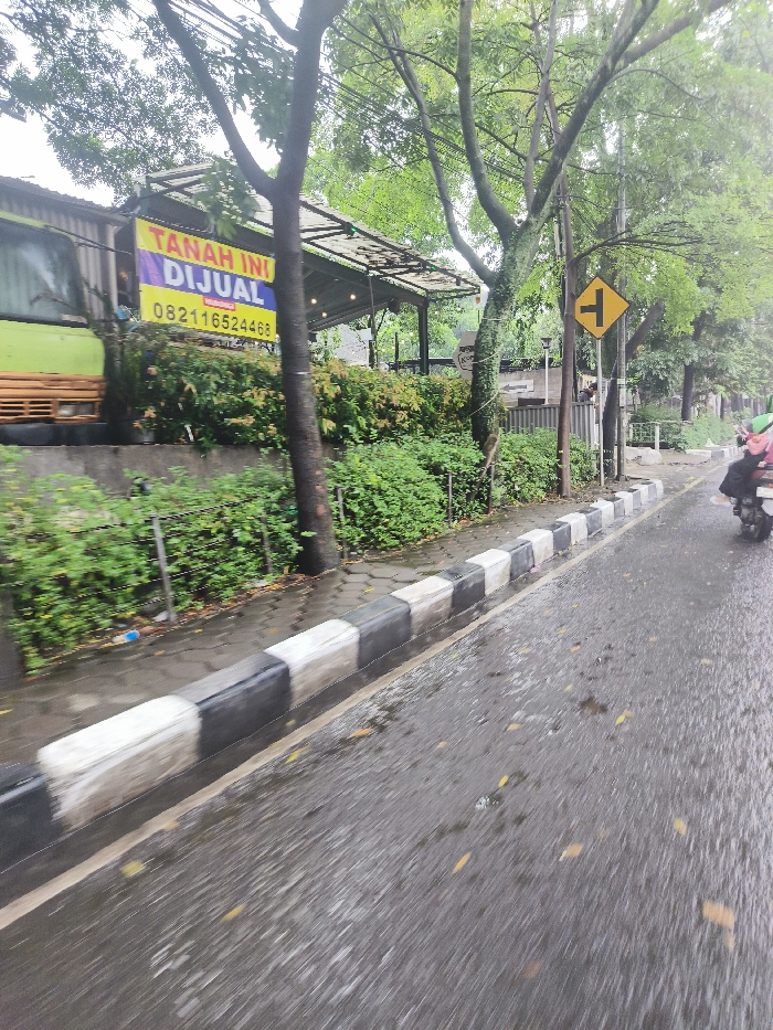 The image shows a wet road in Bandung, Indonesia, with a curb and a sidewalk on the left. A large tree is in the foreground, blocking a view of the road ahead. A yellow sign with a black "T" indicating a T-junction is attached to a post on the right side of the road. A person is driving a motorcycle towards the camera. A sign in Indonesian language advertises land for sale, with a phone number. The road is wet from recent rain, and there is a small amount of vegetation on the side of the road. A large green vehicle is partly visible on the left side of the image. The curb is made of black and white concrete blocks. The image is blurry, likely taken by a person moving or riding in a vehicle.