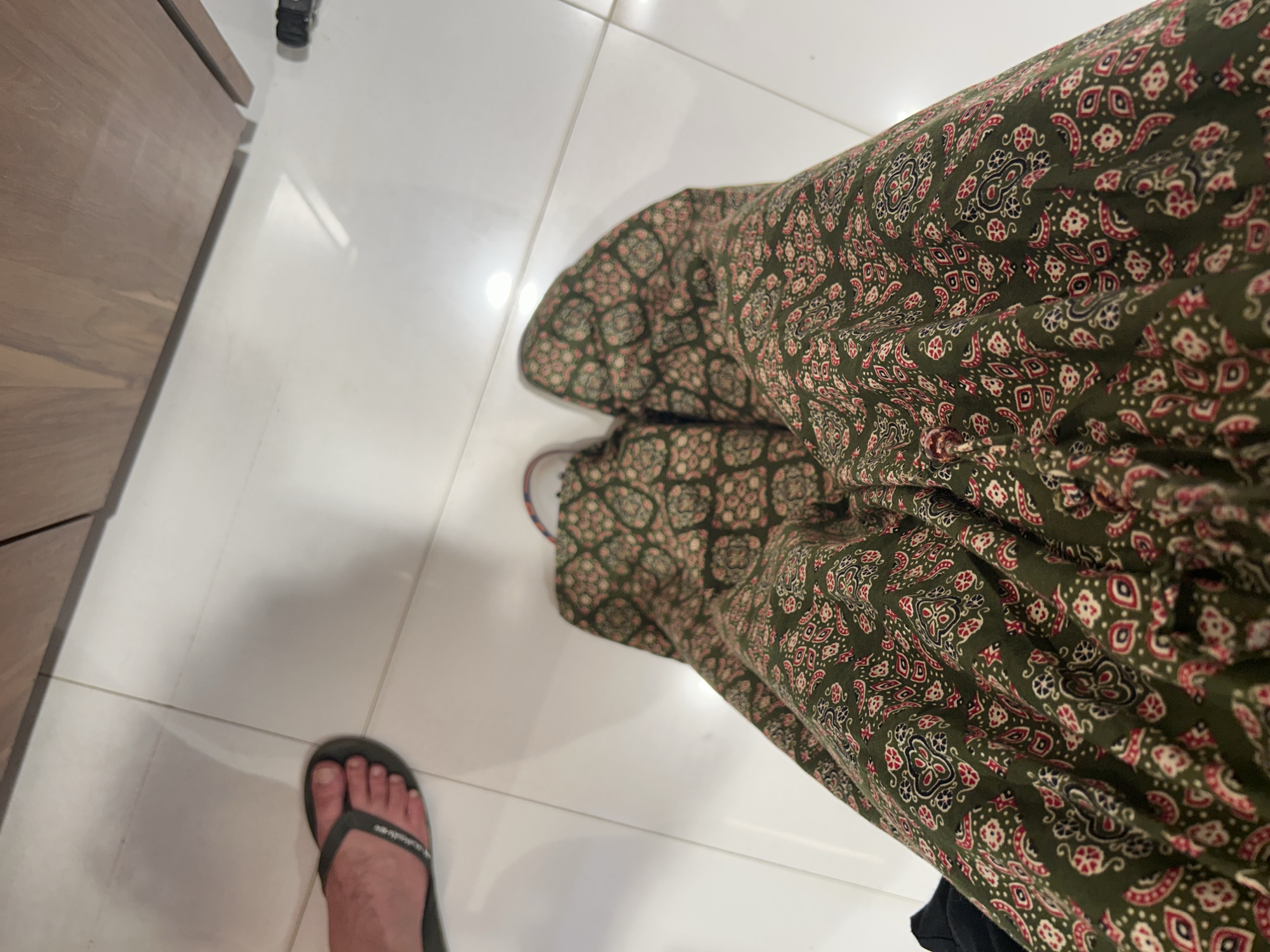 The image shows a first-person perspective of two legs wearing a green patterned skirt and one foot wearing a dark grey flip-flop. The person is standing on a white tiled floor. In the background, a wooden surface is visible, possibly a cabinet or countertop. The scene appears to be indoors, in a residential or commercial setting. There is no visible indication of the location being Mumbai, India.