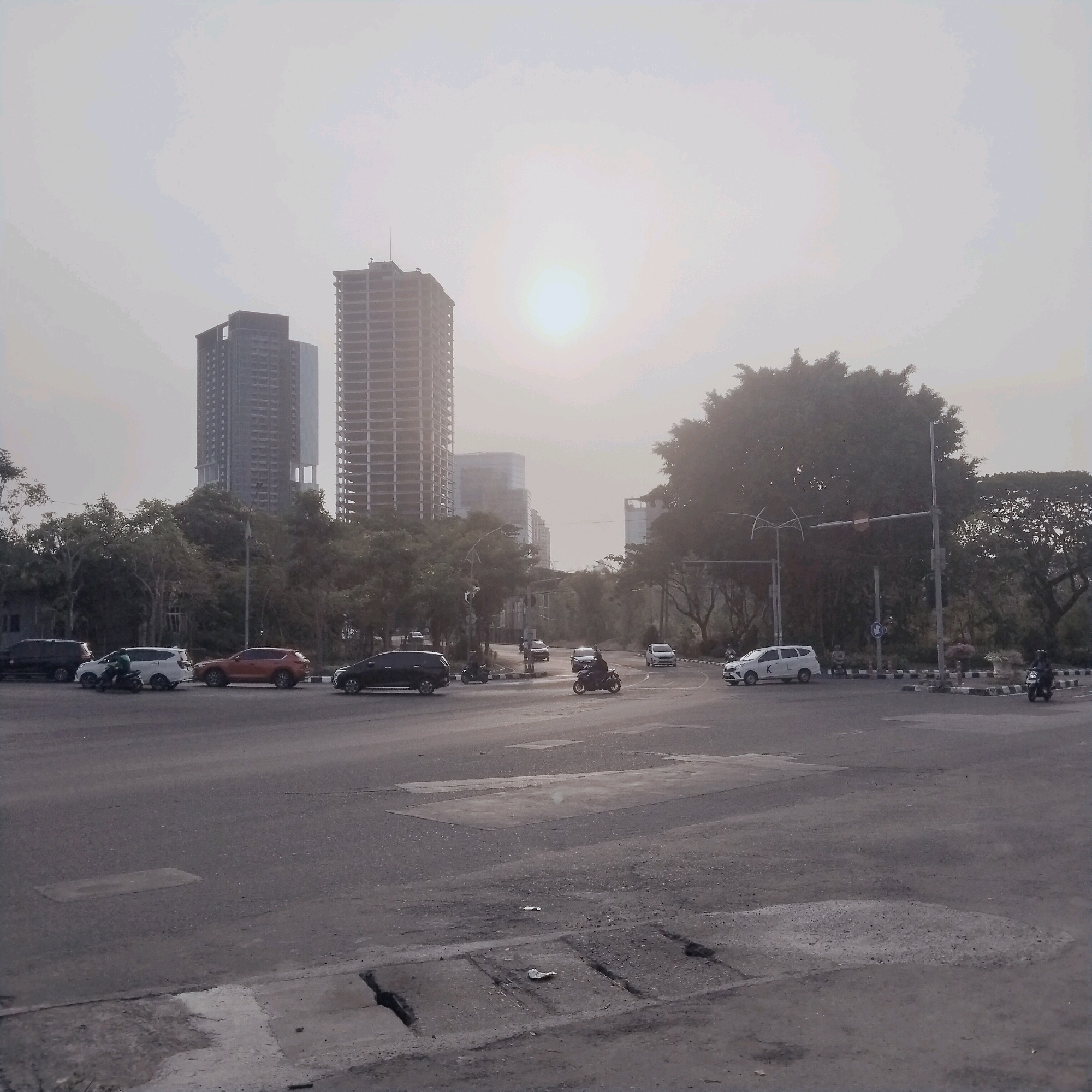 The image depicts a street intersection in Surabaya, Indonesia, under a hazy sky with the sun prominently visible. Several vehicles are present, including cars and motorcycles. On the left, a black SUV is parked, and next to it, a white SUV is moving, with a person on a motorcycle passing by. Further down the road, an orange SUV is visible, followed by a black car. On the right side of the intersection, a white SUV is turning, and a motorcycle rider is proceeding. In the background, tall buildings, some appearing under construction, dominate the skyline. Trees line the sides of the road, and a traffic light is visible. The road surface shows signs of wear, with patches and cracks. The overall atmosphere is urban, with elements of ongoing development.