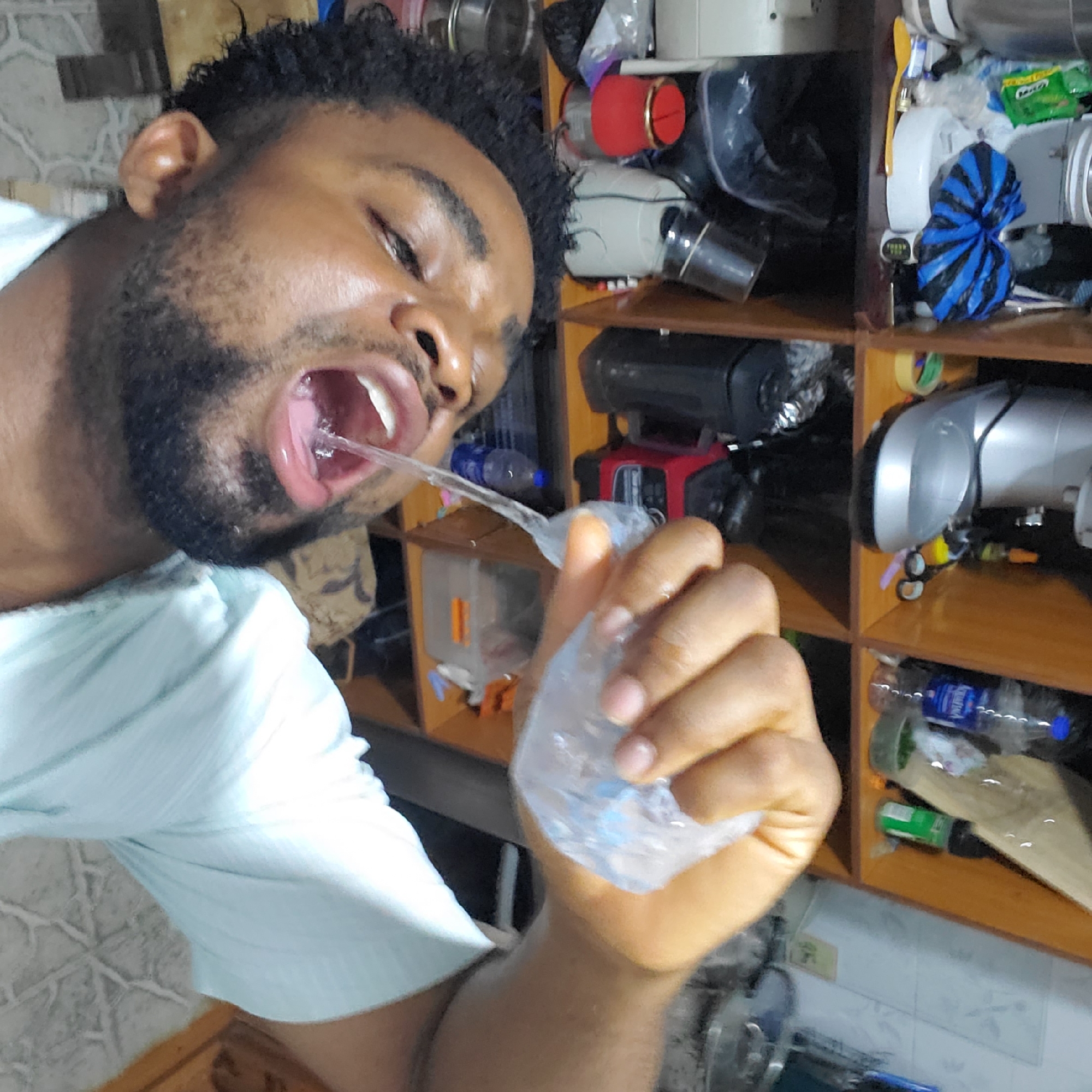 A man with dark hair and a beard is holding a plastic bag of water and squirting the water into his mouth. He is wearing a light blue shirt. Behind him is a wooden shelf filled with various objects, including bottles, containers, and a blue and black striped object. The location appears to be an indoor space, potentially a kitchen or storage area. The city and specific location are Unknown, Unknown.