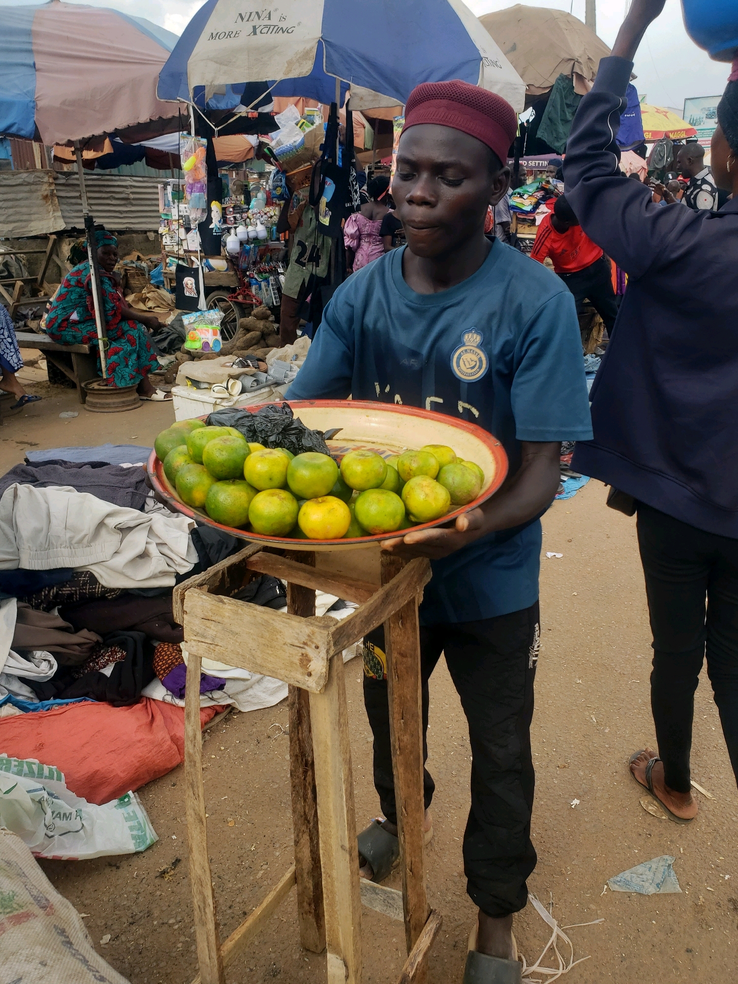 A young man wearing a maroon cap and a blue t-shirt stands in an outdoor market in Sabon Tasha, Nigeria. He holds a large, shallow metal platter filled with green and yellow citrus fruits, possibly oranges. The platter rests on a rustic wooden stand, which is surrounded by piles of clothing and bags. In the background, other market stalls with umbrellas and various goods are visible, along with other people. The ground is a dirt surface, typical of a market setting.