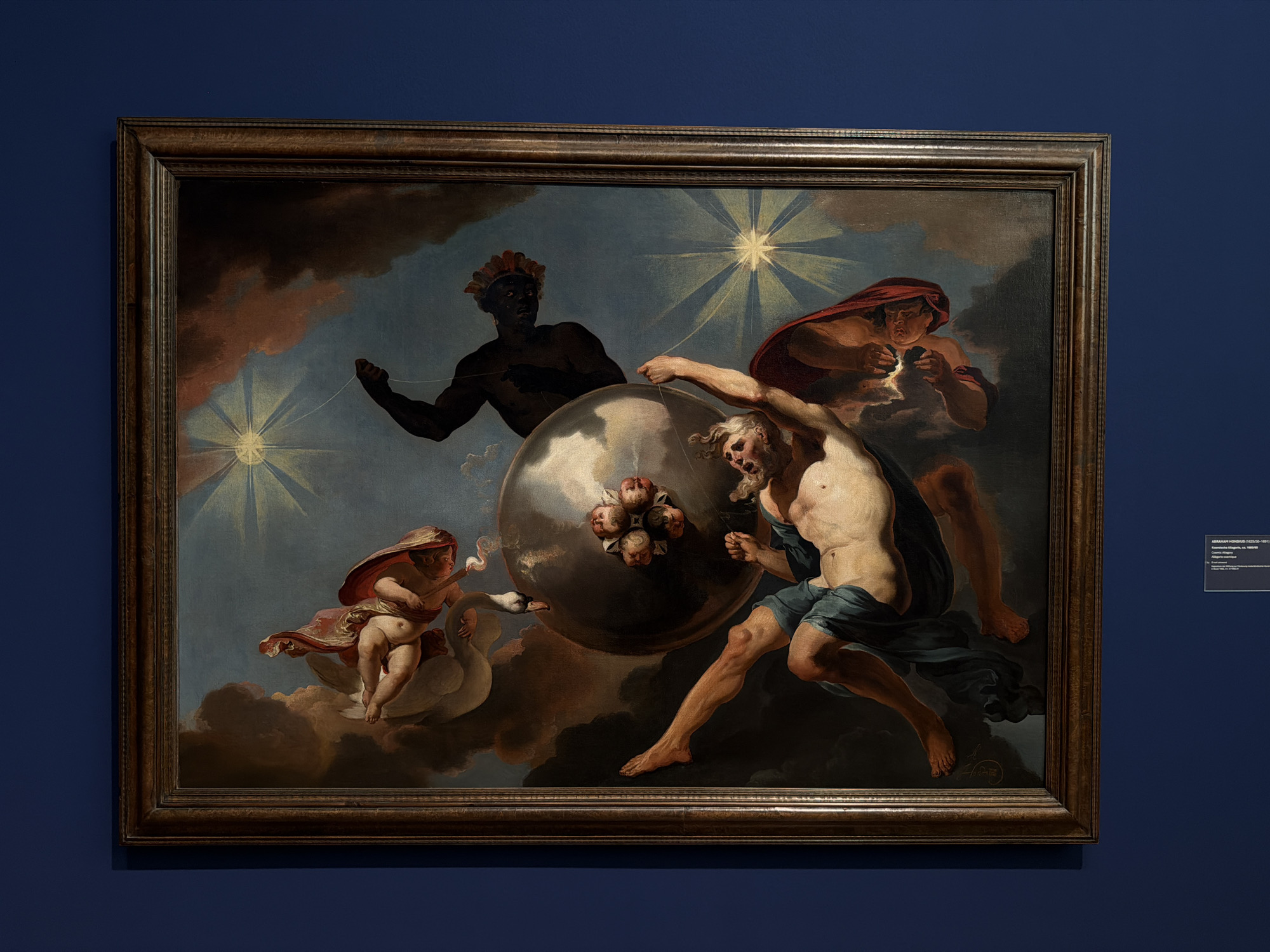 The painting, displayed in a dark wooden frame, depicts a celestial scene with mythological figures. In the center, a large, textured sphere is being manipulated by several beings. An older, muscular male figure with a beard and clad in a blue cloth is pushing against the sphere with his right arm, his body contorted in exertion. To the left, a cherubic figure with wings and a red cloak rides on a white swan, holding a torch that emits smoke. Behind the sphere, a dark-skinned male figure with a headdress made of red feathers is visible, also seemingly interacting with the sphere. Above and to the right, a winged figure with a red cloak is reaching towards the sphere, holding what appears to be a cluster of small, round objects. The background is a twilight sky with prominent starbursts and dark, swirling clouds, suggesting a cosmic or divine setting. The painting is hung on a deep blue wall. To the right of the painting, a small white label is affixed to the wall, providing information about the artwork, including the artist Abraham Hondius (1628-1691) and the title "De overwinning van de tijd op de skulptuur" (The Victory of Time over Sculpture). This artwork is located in the Kunstmuseum Basel, Switzerland.