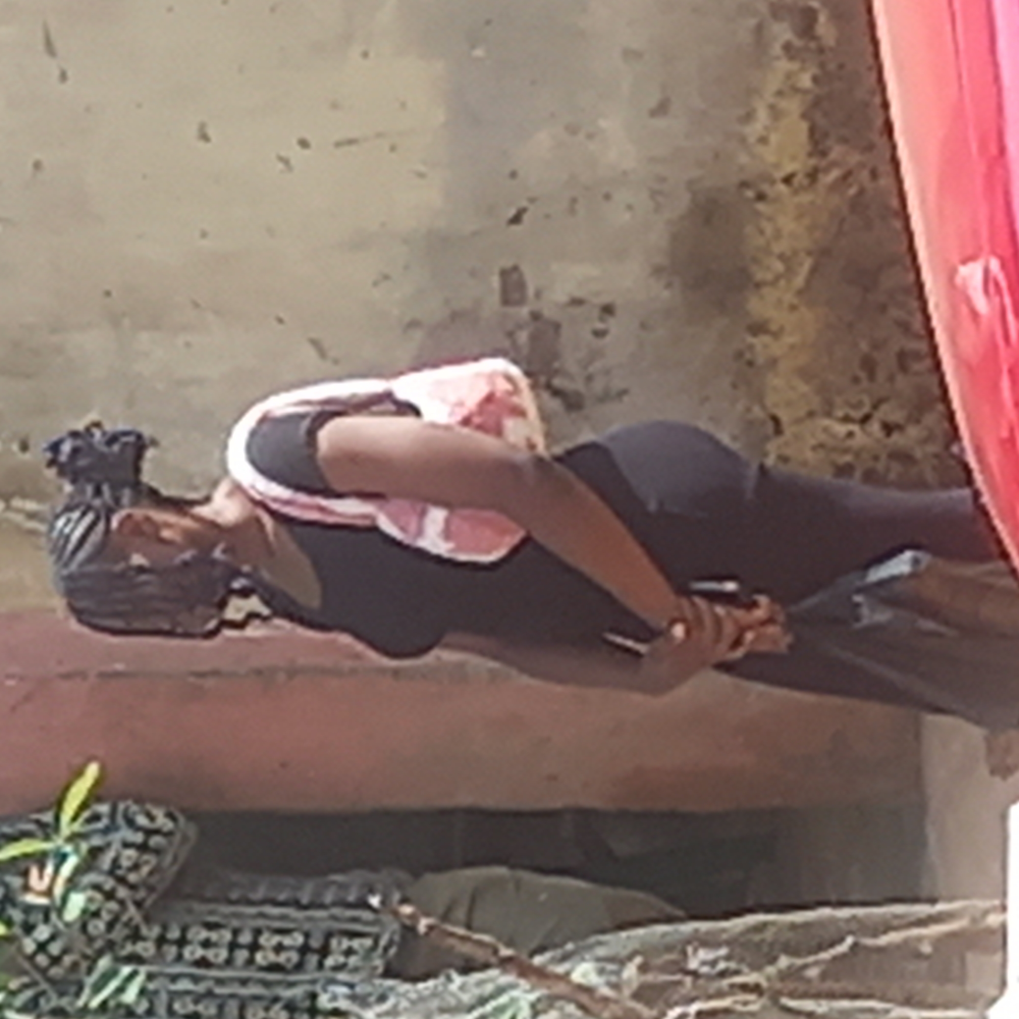 A woman with braids and a pink and white patterned backpack is outdoors in what appears to be Dadi Riba, Nigeria. She is wearing a black shirt and black leggings, and is holding what appears to be a cell phone. She is looking down at her phone, and there is a red car parked to the right. The background is a concrete wall, and there are some plants and debris on the ground.