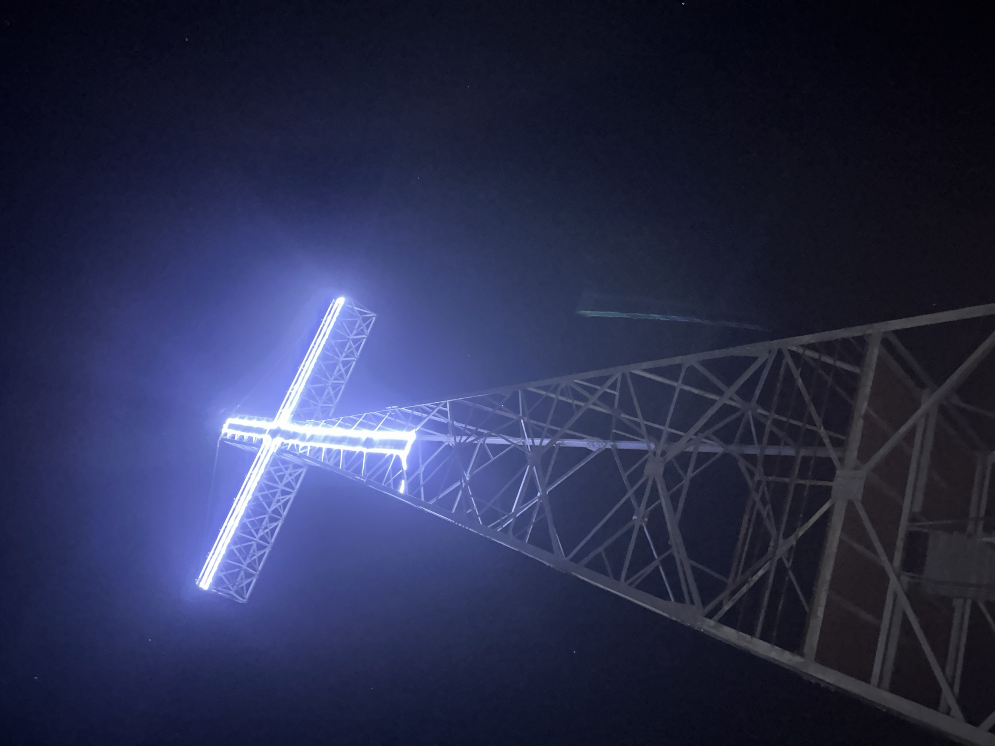 View looking up at a tall steel structure supporting a large illuminated cross on the city of Unknown, Unknown, at night. The cross glows with a soft blue light, contrasting sharply with the darkness surrounding it. The steel structure is intricate, with many intersecting beams and supports visible against the night sky. A few faint stars are visible in the upper part of the frame.