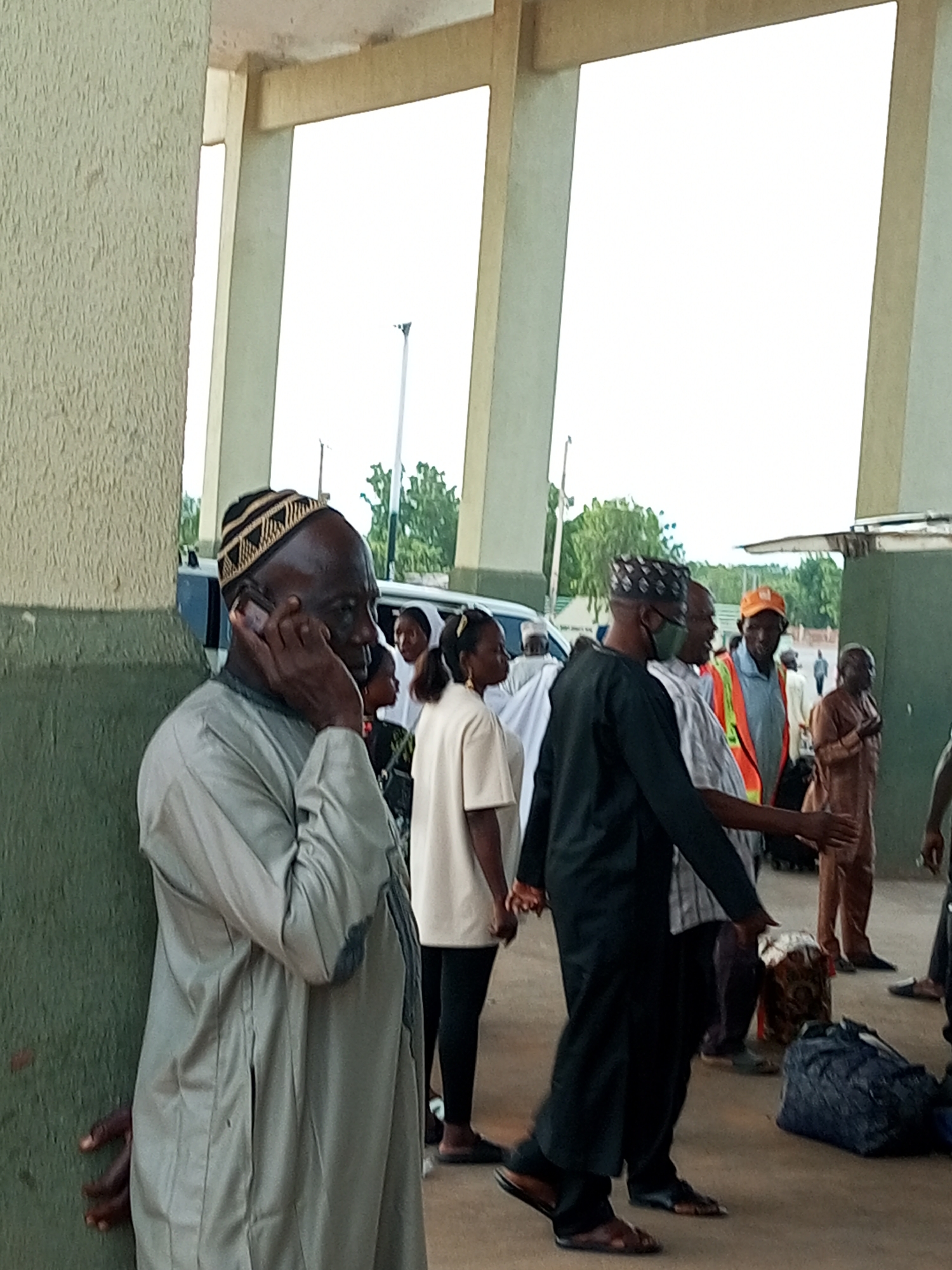 In Gombe, Nigeria, a group of people stand near a building with a textured green wall and exposed concrete pillars. A man in a grey traditional outfit and a patterned cap is on his phone, leaning against the green wall. To his right, a woman in a white outfit and black leggings stands looking towards the right. Further back, a man in a black traditional outfit and a face mask is interacting with another man wearing an orange cap and a reflective vest. Luggage, including a blue duffel bag and a patterned bundle, is scattered on the ground. The environment appears to be an outdoor waiting area, possibly a transport terminal, with glimpses of trees and a bright sky in the background.
