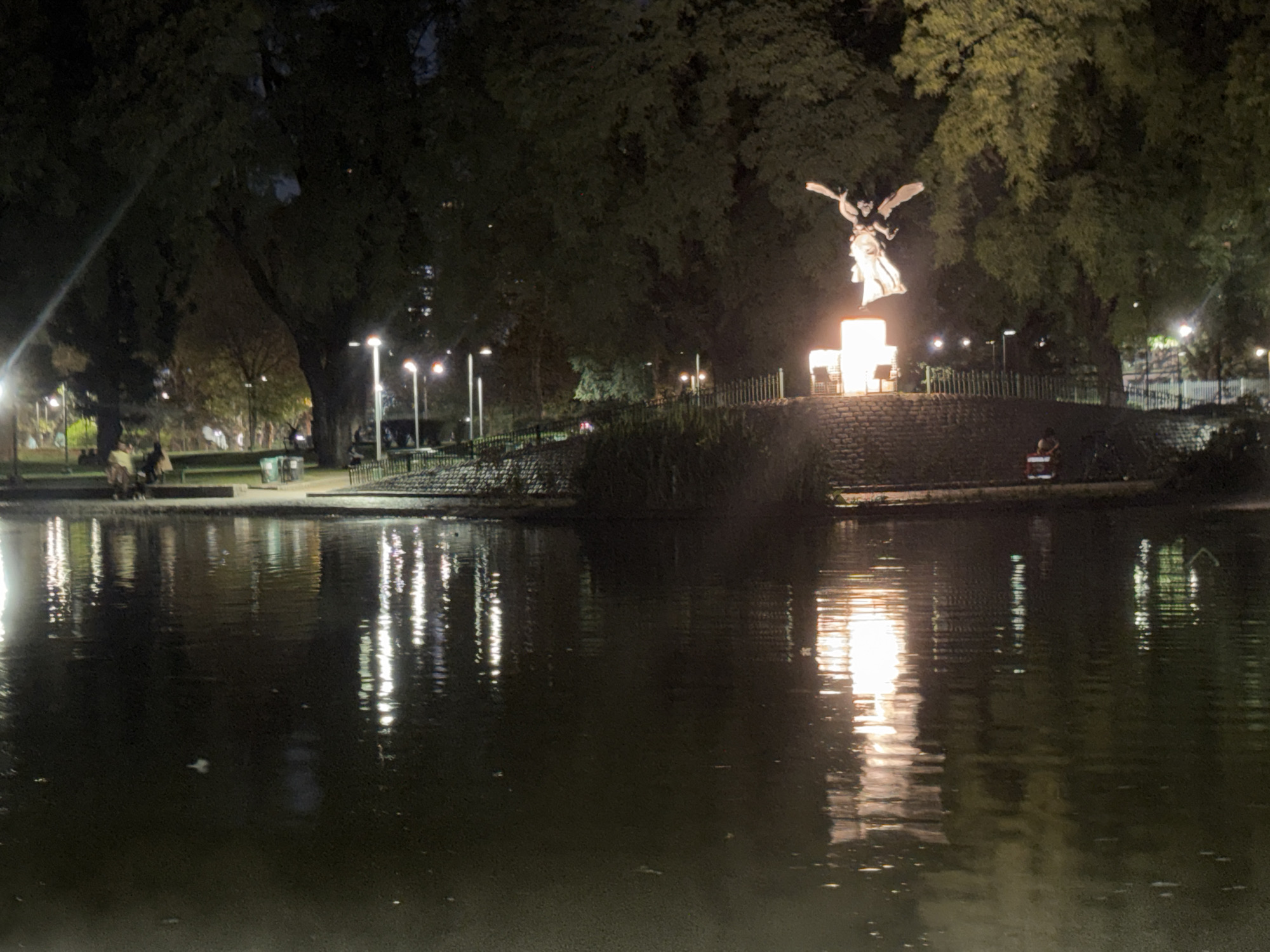 The image shows a nighttime view of a lake in Parque 3 de Febrero (also known as Bosques de Palermo), a large park in Buenos Aires, Argentina. The lake reflects the lights and some of the surrounding environment. In the center of the image, a brightly lit statue of an angel stands on a raised platform near the edge of the lake, surrounded by trees. To the left of the statue, there are people sitting on benches near the water's edge. In the right side of the frame, a person sits by the lake. Lights are visible along the perimeter of the park. The water appears calm, with reflections of the surrounding lights creating a textured surface. The overall scene is dimly lit and evokes a sense of tranquility.