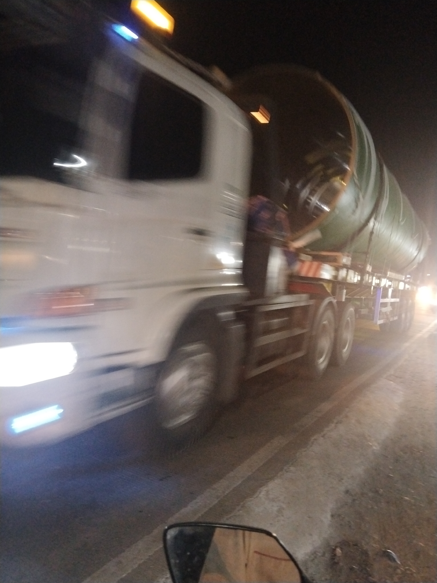The image shows a blurry, motion-blurred view of a white tanker truck at night in the city of Suka Ramai, Indonesia. The truck is driving on a road, and its lights are on. The tanker is large and green. The scene appears to be a street or highway with some reflective markings on the road. A side mirror from what appears to be another vehicle is visible in the foreground. The overall impression is of a vehicle moving at speed at night.