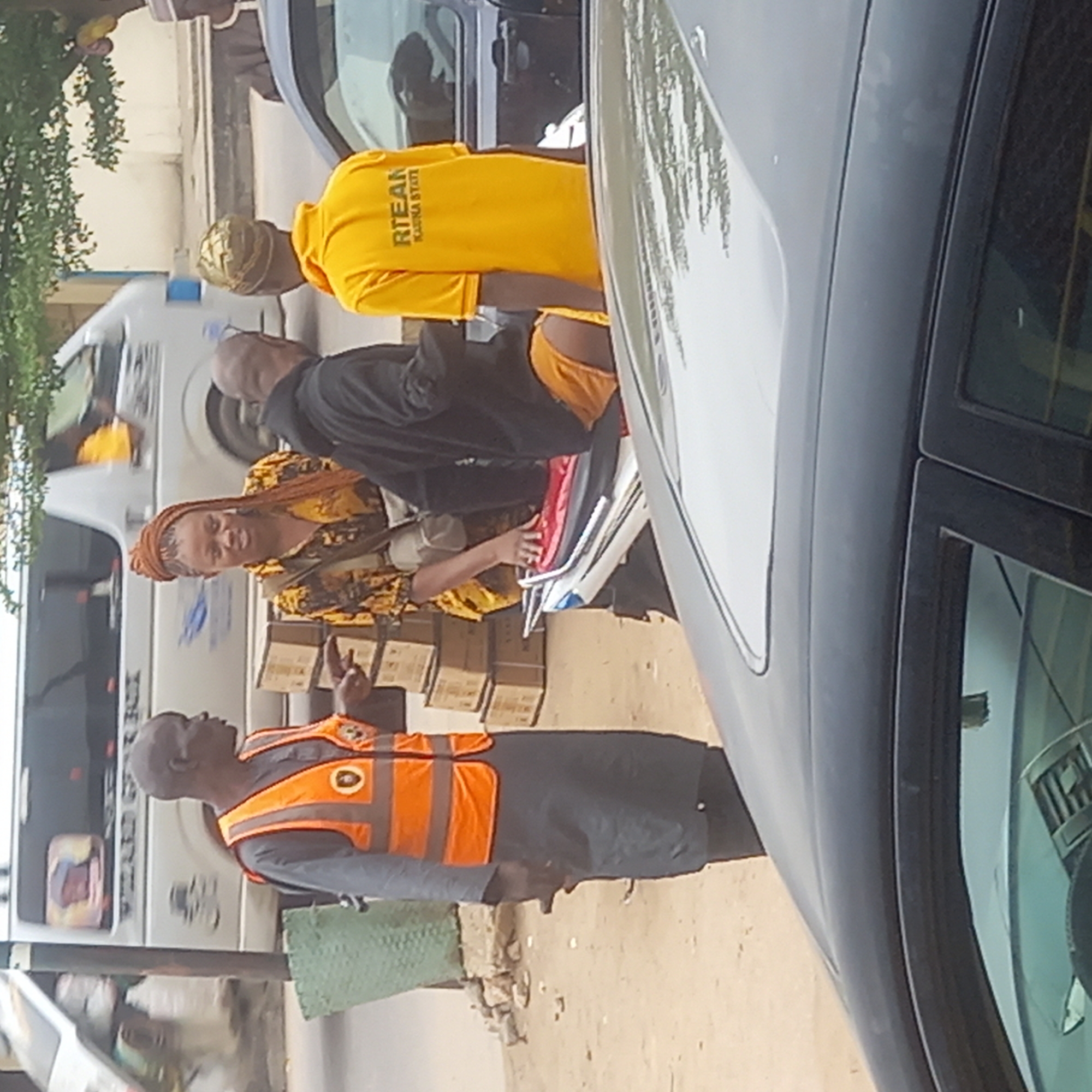 The image depicts a street scene in Dadi Riba, Nigeria. Several people are visible, including a man wearing an orange traffic vest, a woman with a headwrap and patterned yellow clothing, a man in a black shirt, and a person in a yellow shirt with "RTEAN KADUNA STATE" printed on the back. The people are gathered near several vehicles, including buses and cars. Boxes are visible near the woman, and papers are being handled by someone near the yellow-shirted person. The environment appears to be an outdoor street setting with trees in the background.