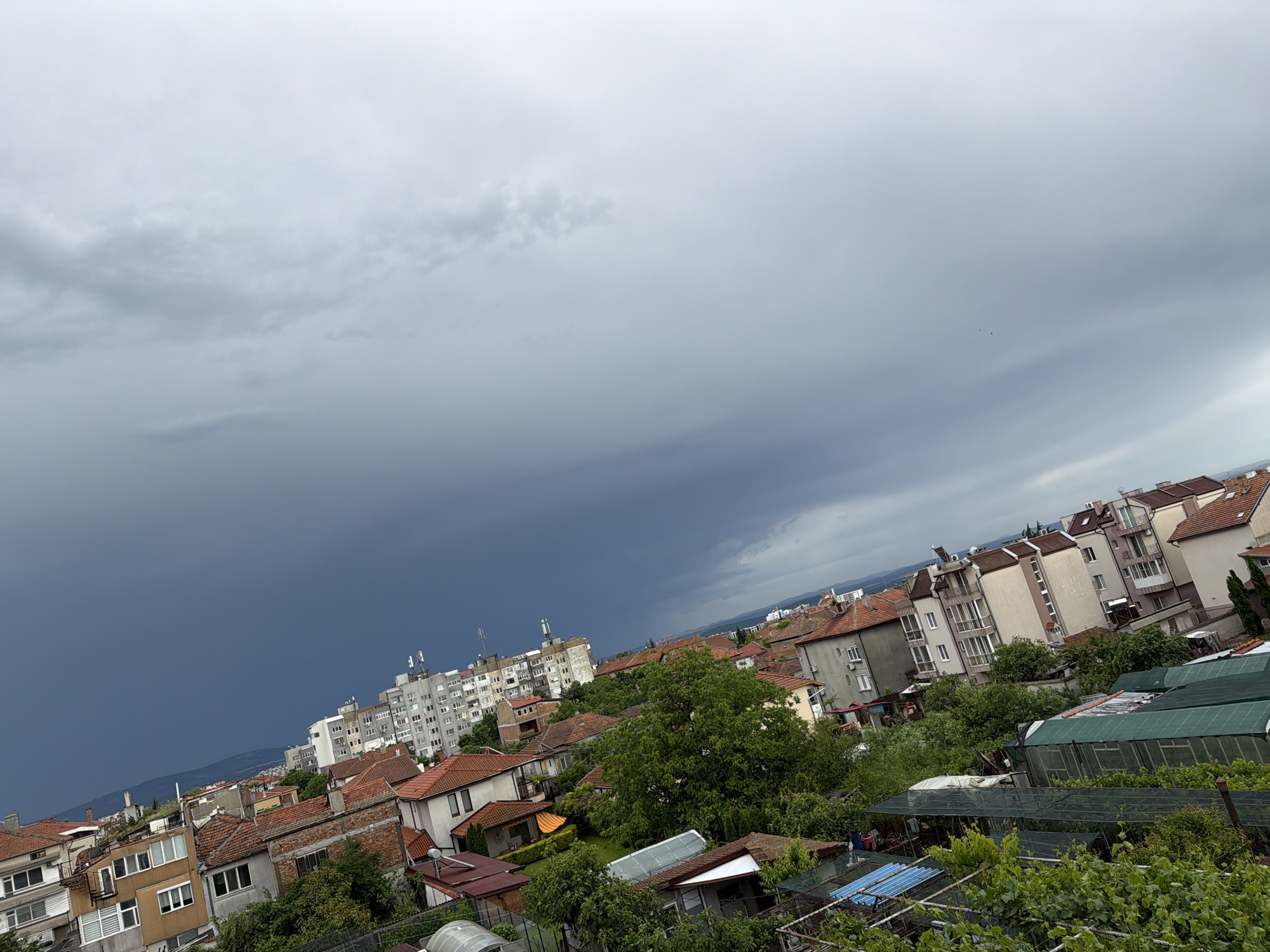 The image presents an oblique, elevated view of a residential area in Burgas, Bulgaria, taken during a stormy day. In the foreground and midground, houses with red tile roofs intermingle with denser apartment buildings. A patch of dark, ominous storm clouds fills a portion of the sky, suggesting an approaching downpour. The overall lighting is dim due to the storm clouds. Vegetation is visible in gardens and between buildings, adding green tones to the scene. Some distant mountains are visible in the far background.