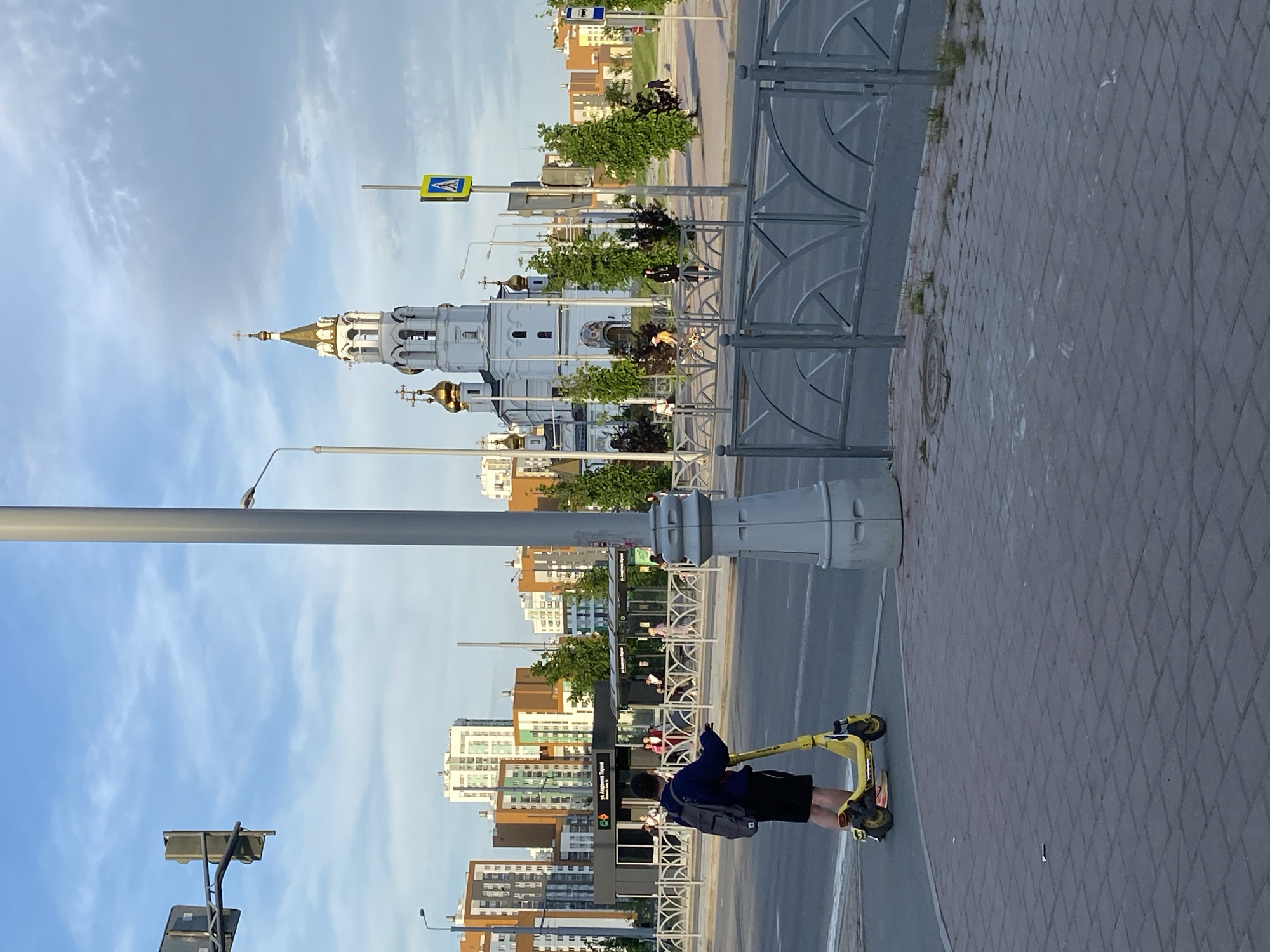 The image shows a cityscape in Yekaterinburg, Russia. A person is riding a yellow electric scooter on a brick-paved sidewalk. A gray metal fence runs parallel to the sidewalk, separating it from a multi-lane road with light traffic. Across the road is a row of modern apartment buildings and a bus stop/shelter. In the background, partially obscured by trees and streetlights, stands a white church with golden domes and a steeple. The sky is blue with scattered clouds. There are traffic lights, pedestrian crossings, and street signs visible.