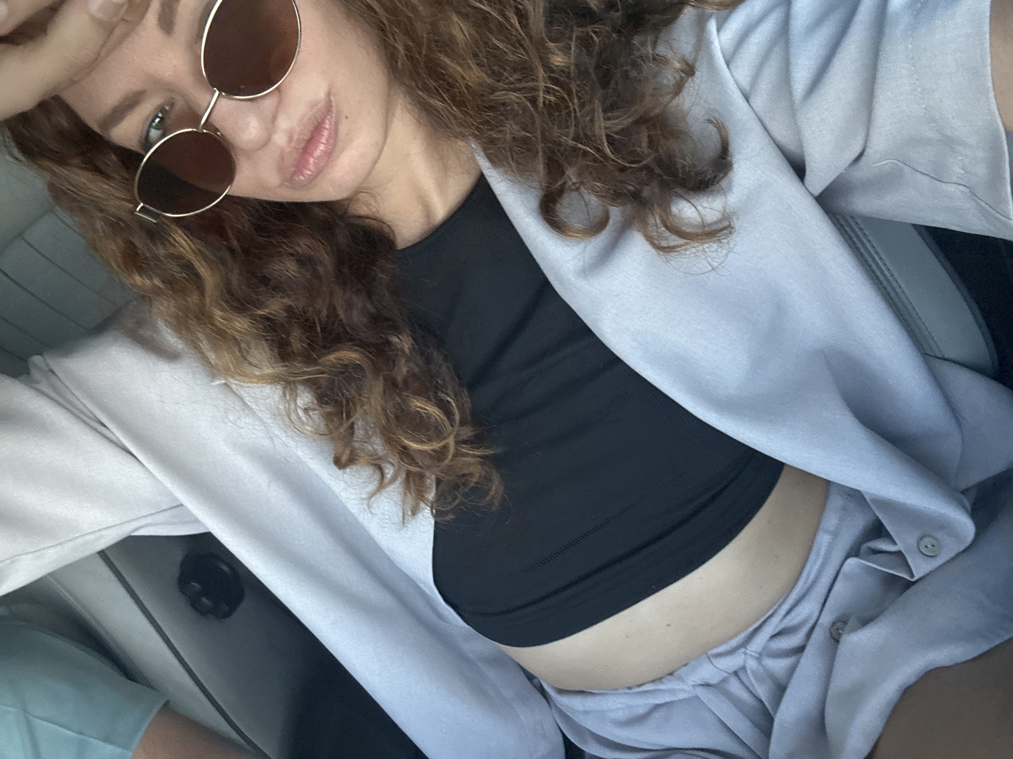 A woman with curly brown hair and sunglasses is in a car. She is wearing a black crop top and a light-colored blazer. The background shows a car interior with seats and a vent. The overall setting appears to be daytime.