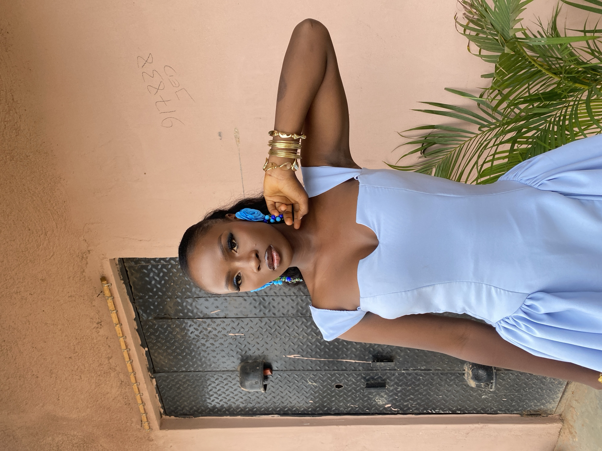 This media file features a young Black woman standing outdoors. She is wearing a light blue dress with ruffled details on the skirt and off-the-shoulder sleeves. She accessorizes with multiple gold bracelets on her left arm and large blue floral earrings. Her makeup is visible, with dark eyeliner and a glossy lip. Her left arm is bent, with her hand resting on her head.
The background consists of a textured, light pink wall with some graffiti, and a metal door or gate with a diamond plate pattern. To the right of the woman, palm fronds from a plant are visible. The lighting suggests it is daytime, likely outdoors. The overall mood appears to be a casual portrait or fashion shot.
Visible text on the wall includes "917838" and "027". Based on the provided location context, the setting is Gwong, Nigeria.