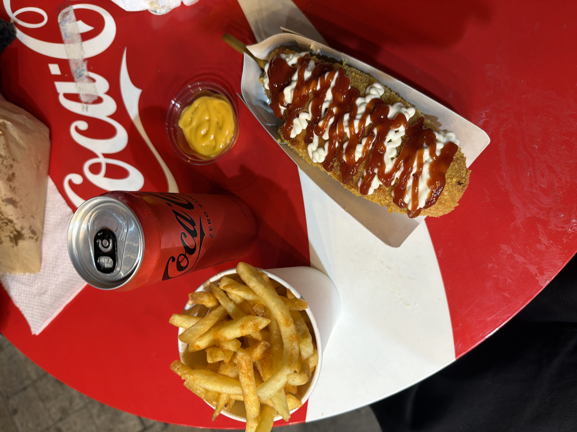 An overhead, close-up shot captures a Coca-Cola themed table featuring a can of Coca-Cola Zero Sugar, a cup of french fries, a small container of yellow mustard, and a corn dog drizzled with ketchup and mayonnaise. The table's surface is predominantly red with white accents, and a white paper napkin is visible in the upper background. The corn dog is served in a white paper tray, and the french fries are in a white paper cup. The scene appears to be set outdoors, possibly at a food stand or casual dining establishment. The lighting suggests it is daytime. The location is in Belgrade, Serbia.