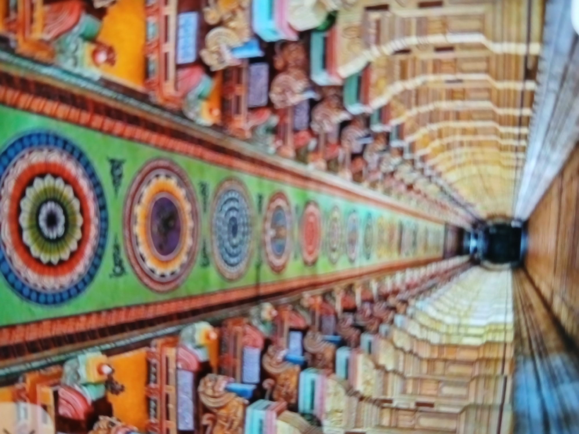 The image depicts the interior of a hall in a temple, likely located in Bengaluru, India. The walls are adorned with colorful circular patterns and sculptural figures. The ceilings appear to be carved and ornamented, creating a symmetrical and receding perspective. The hall seems long, leading to a dark area in the distance. There are no people visible. The environment suggests a place of worship with traditional Indian artistry.