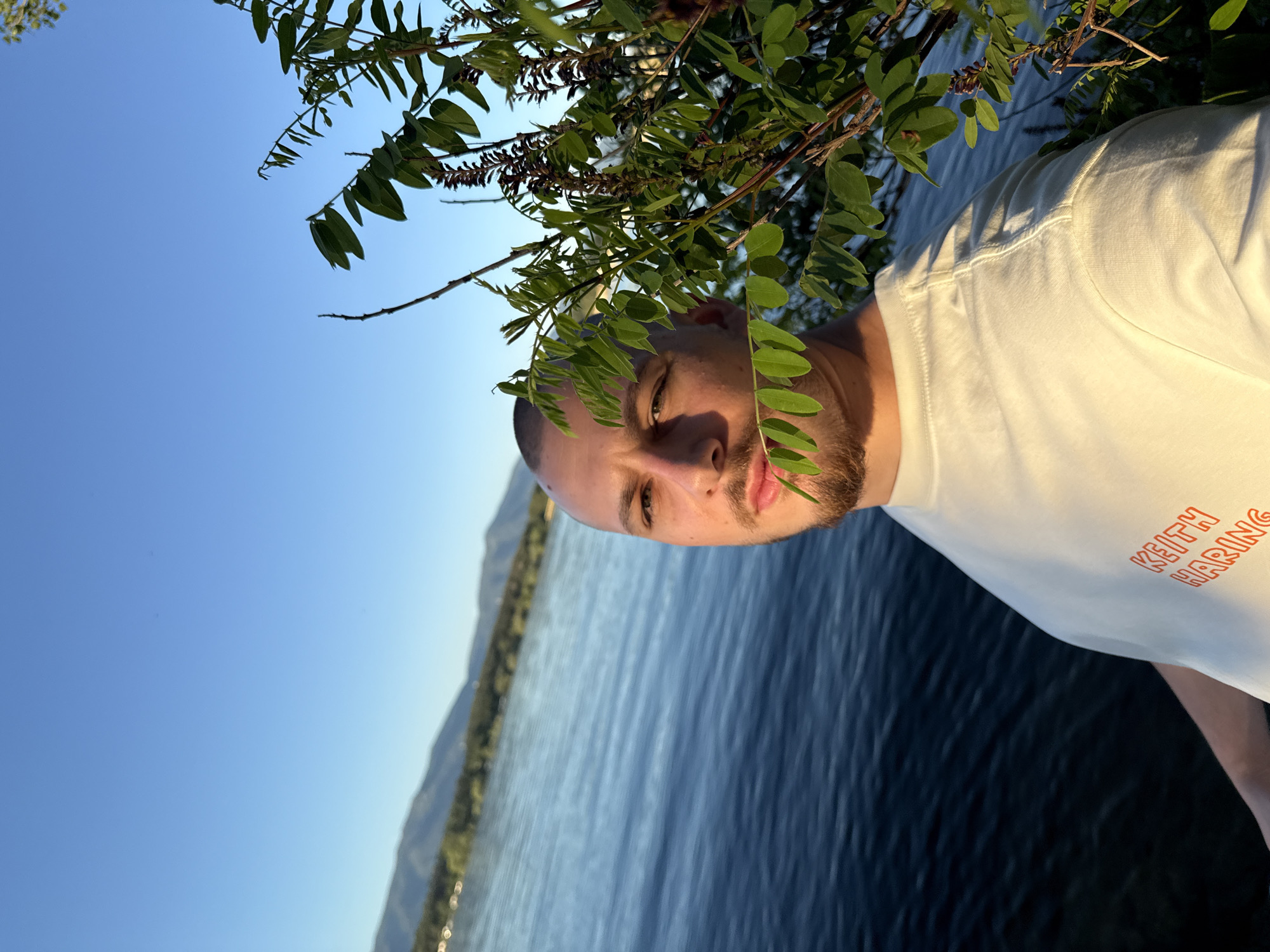 A bald man with a beard is photographed outdoors near the city of Koprinka, Bulgaria. He is wearing a white t-shirt with "KEITH HARING" printed in orange on the left side. The man is looking towards the camera, with green leaves from a plant partially obscuring his face. In the background, a large body of water stretches out, with a mountainous landscape visible on the opposite shore. The sky is clear and blue, illuminated by what appears to be the golden hour light. The man is positioned on the left side of the frame, with the plant on the right, creating a sense of depth. The overall scene conveys a peaceful and natural setting.