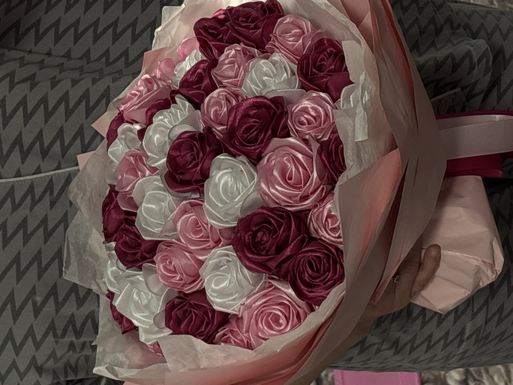 The image depicts a bouquet of artificial roses, primarily in shades of white, light pink, and deep burgundy, nestled in white tissue paper and wrapped with a pink outer layer and ribbon. The bouquet is being held by a hand adorned with a ring. The background features a grey and white chevron-patterned fabric. The location is not determinable from the image.