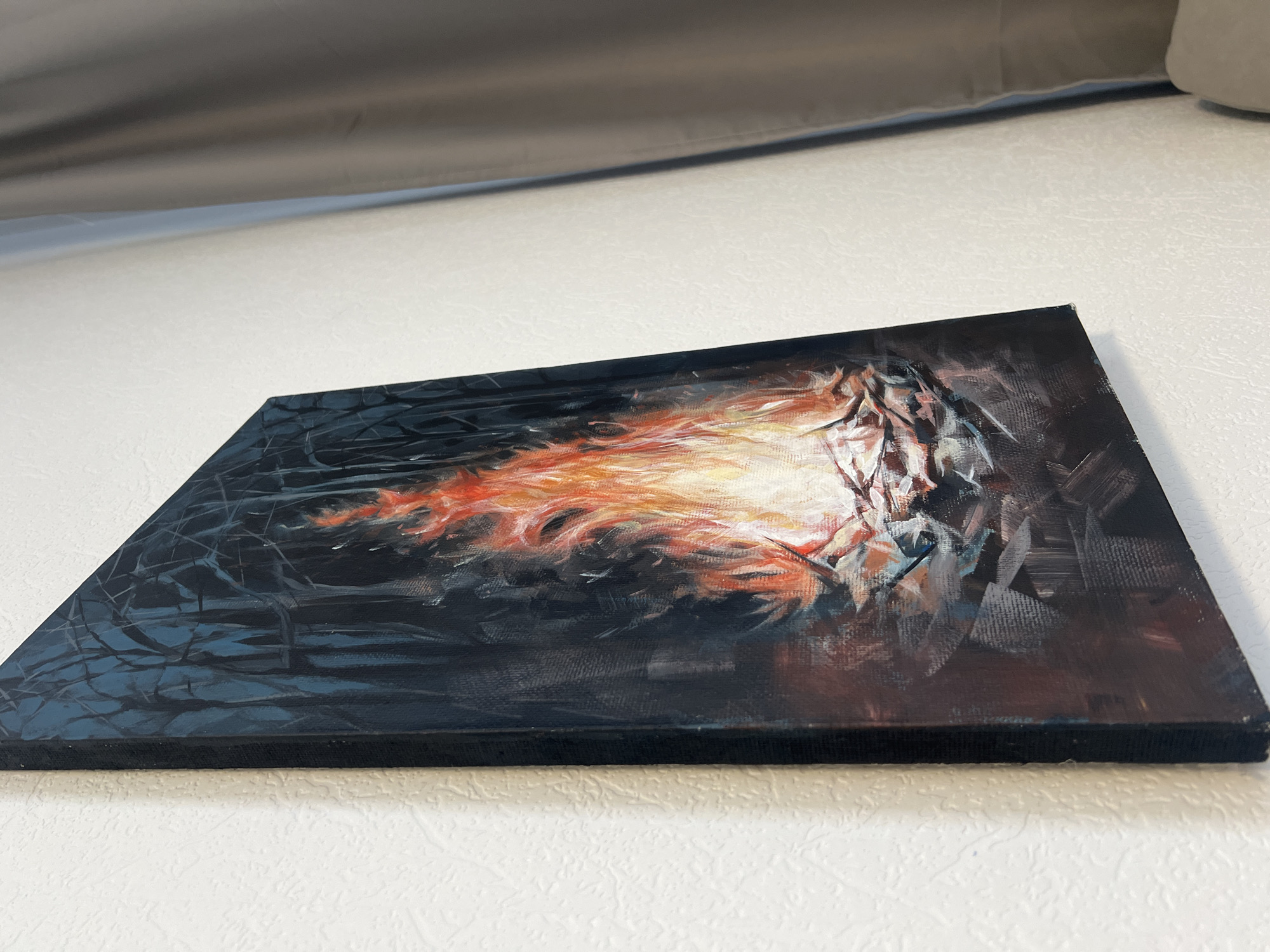 A rectangular canvas painting is on a white surface, likely a tabletop. The artwork features a dark background with a central element of orange and white, possibly depicting fire or a fiery creature. Dark lines and brushstrokes extend from the central element, suggesting a forest or cave setting. The canvas is stretched over a wooden frame. The image's depth of field is shallow, and the lighting is moderate. A light grey object is behind the painting. There is no indication of this being in the city of Tyumen, Russia.