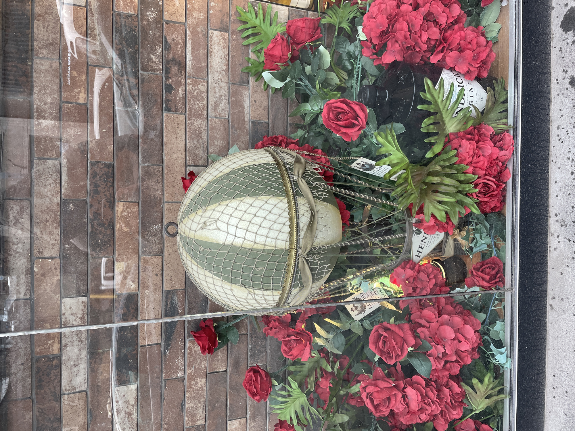 The image displays a retail shop window in Yerevan, Armenia, showcasing a decorative arrangement. Centrally featured is a cream-colored hot air balloon model with vertical green stripes, a white net overlay, and a small, open-weave basket suspended beneath it. This model is embedded within a dense arrangement of artificial red roses and clusters of smaller red flowers, accented by green foliage, including broad, lobed leaves. Two dark-colored bottles are visible among the flowers; at least one is clearly identifiable by its white label as "HENDRICK'S GIN." Small decorative tags, one bearing the word "LOVED" and additional text, are also present. The interior background of the display consists of a multi-toned brick wall. The clear glass storefront reflects elements from the exterior, including faint, reversed text that appears to read "BANK OF ARMENIA." The immediate foreground shows a grey concrete sidewalk. No individuals are depicted.