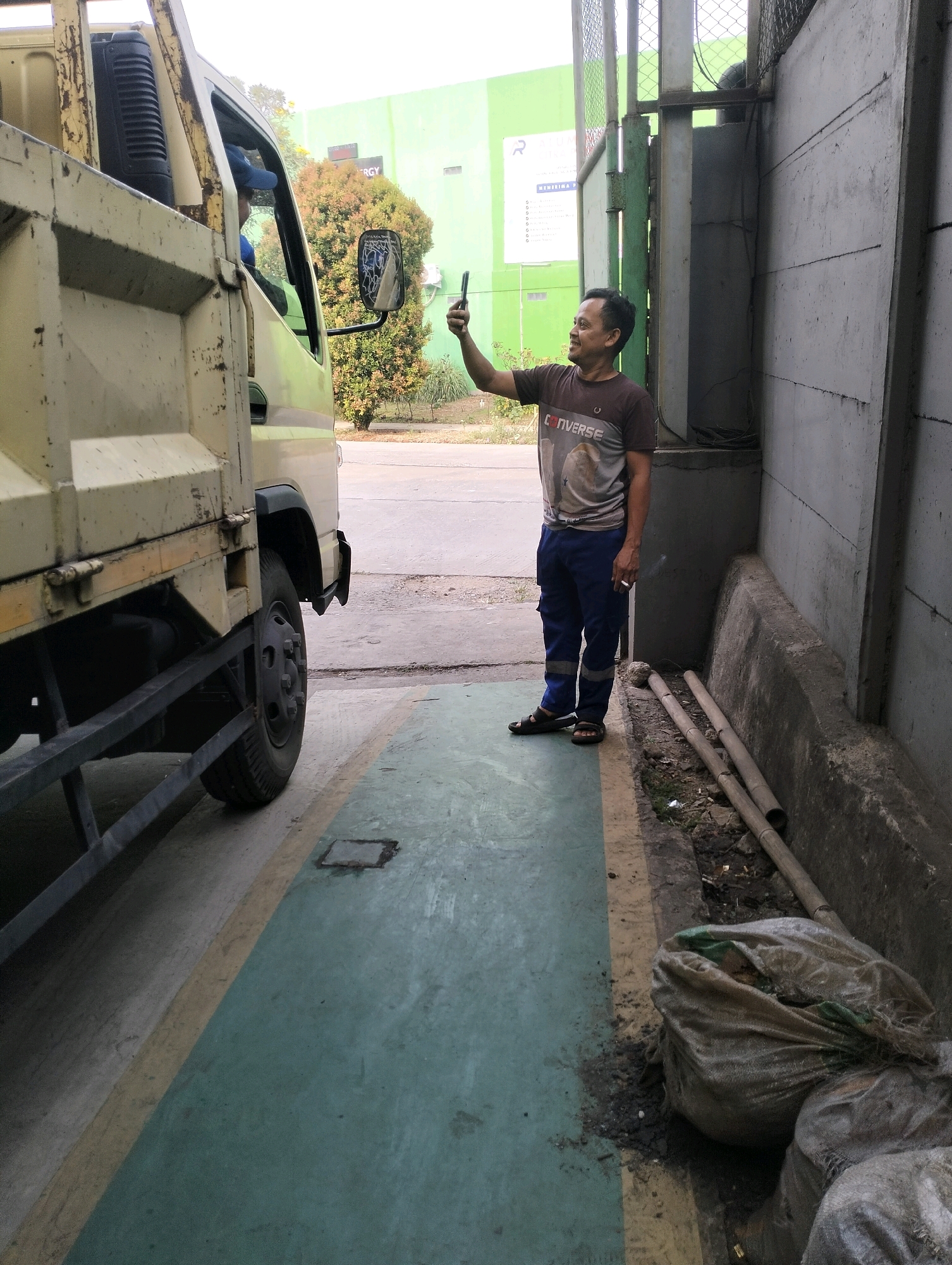 A man is standing and holding a smartphone to take a picture or video. He is wearing a brown t-shirt with "Converse" printed on it, blue pants, and black sandals. He has a cigarette in his right hand. He is standing next to a light-colored dump truck parked on a paved area with a green and yellow painted lane. Behind the man is a concrete wall. To the right of the man and truck, there are two large bags filled with what appears to be trash, and some bamboo poles. In the background, a building with a green facade and a sign is visible, along with trees. The scene appears to be an industrial or commercial area in Gunung Sindur, Indonesia.