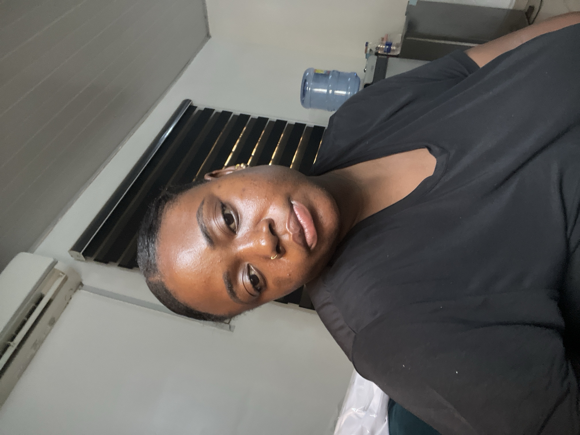 A medium indoor shot depicts a dark-skinned woman with a nose ring wearing a black shirt. An air conditioning unit is visible on the lower left. In the background, there is a water dispenser on top of a gray surface and window blinds. The location is consistent with a room in Abuja, Nigeria.