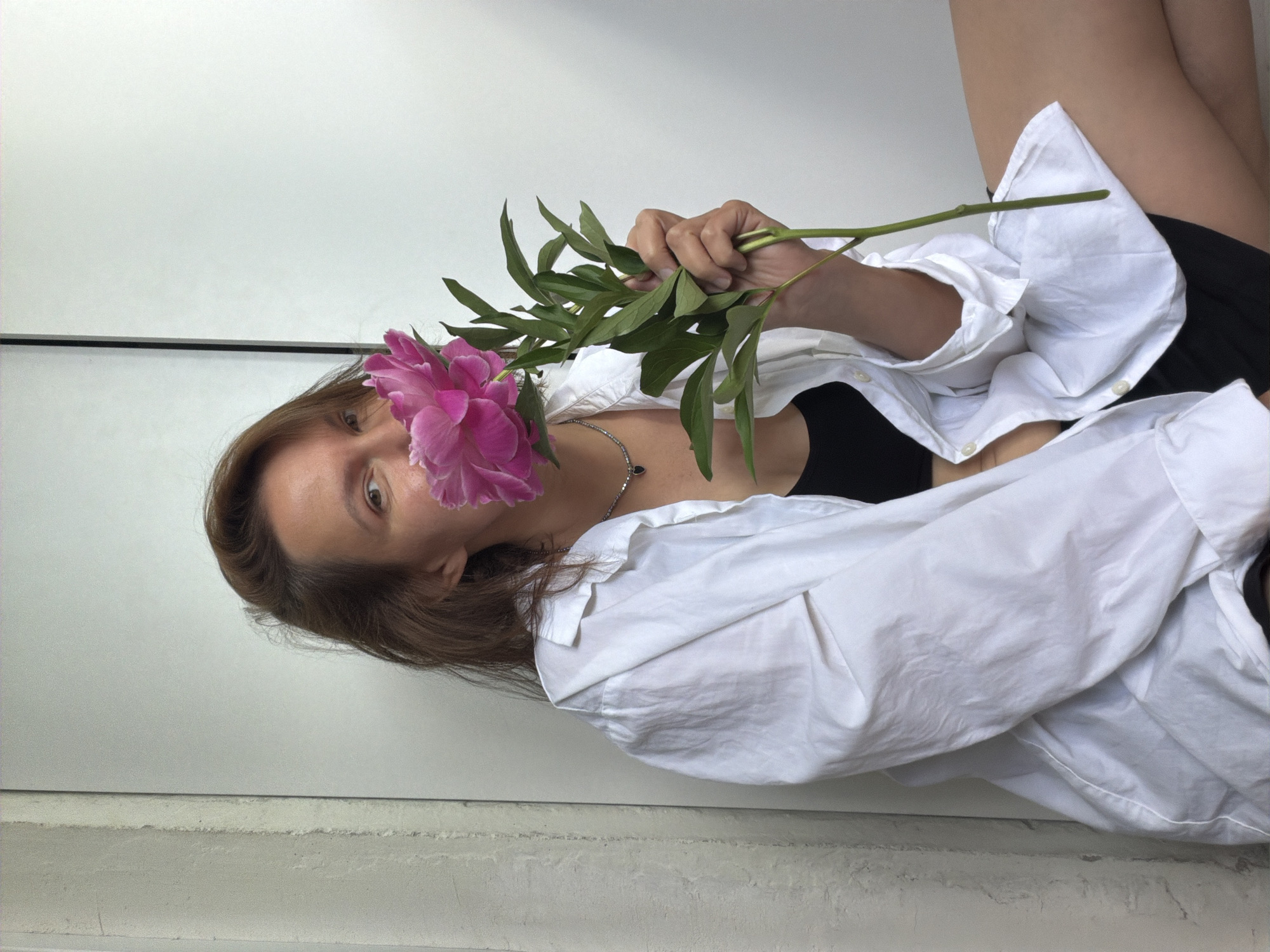 A young woman with brown hair is sitting on the floor, wearing an unbuttoned white shirt over a black top and black shorts. She is holding a pink peony in front of her face, with her eyes visible over the flower. The background appears to be an indoor setting with a white wall and a narrow vertical partition on the left. The lighting is soft and natural. There are no specific landmarks or features that would allow for identification of the location as Chaikovsky, Russia.