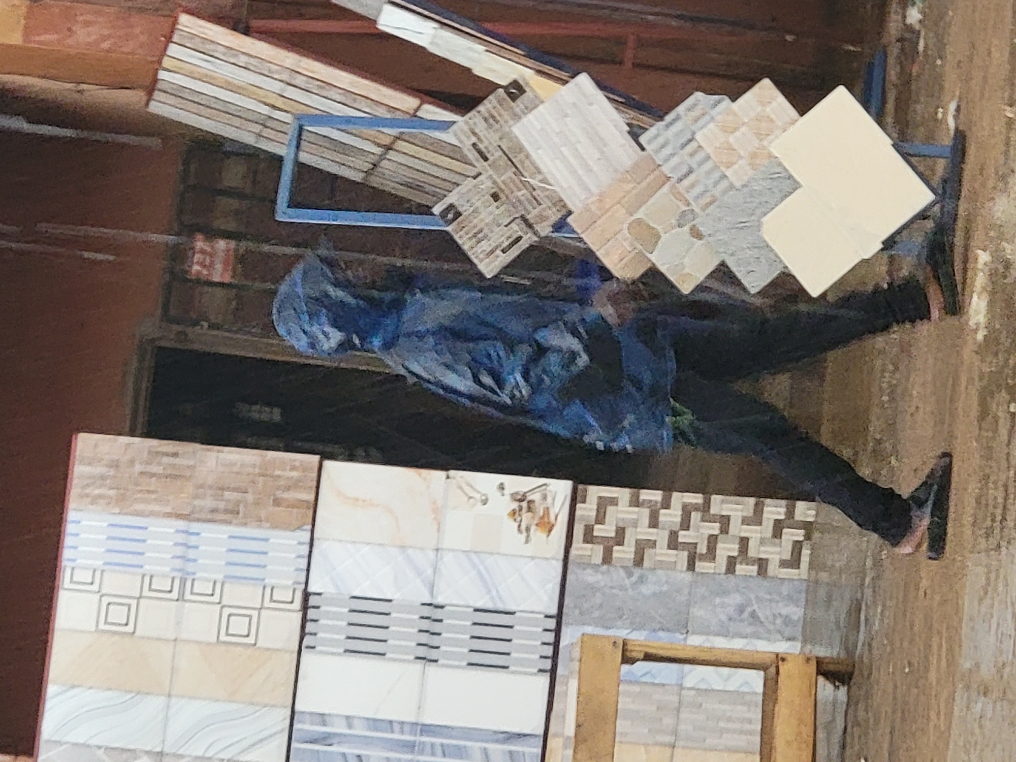 This vertically oriented image, captured in Maiburiji, Nigeria, depicts a person standing amidst a display of floor and wall tiles, likely within a building material store or an outdoor market stall.
The primary subject is a person, whose face is obscured by a dark blue hood, wearing a matching dark blue jacket or raincoat and dark pants. They are standing with their left leg slightly forward, facing towards the right side of the frame, seemingly observing the tile displays.
The scene is dominated by various ceramic or porcelain tiles. In the background, a series of smaller, patterned tiles are arranged on an angled blue metal display rack. These include tiles with intricate geometric designs, stone-like textures, and plain light colors. In the foreground, larger rectangular tiles are displayed vertically, showcasing a diverse range of patterns, including brick-like textures, horizontal stripes, abstract waves, and geometric squares.
The setting appears to be a practical, possibly open-air or semi-covered commercial space. The ground is a light, dusty brown. In the upper background, construction materials like wooden planks are visible, alongside what appears to be a blue tarp or bag. A dark doorway or opening is situated behind the person. A rough, brick-like wall section is also discernible in the background.
There is no specific activity evident beyond the person standing and observing. The time of day appears to be daytime, possibly overcast or shaded, as there are no harsh shadows. No distinct weather conditions can be inferred, though the hooded attire might suggest protection from elements.
A small, blurry red and white object with unreadable text is visible on a background wall section, but no other readable text is apparent. The overall impression is that of a bustling commercial environment focused on building supplies.