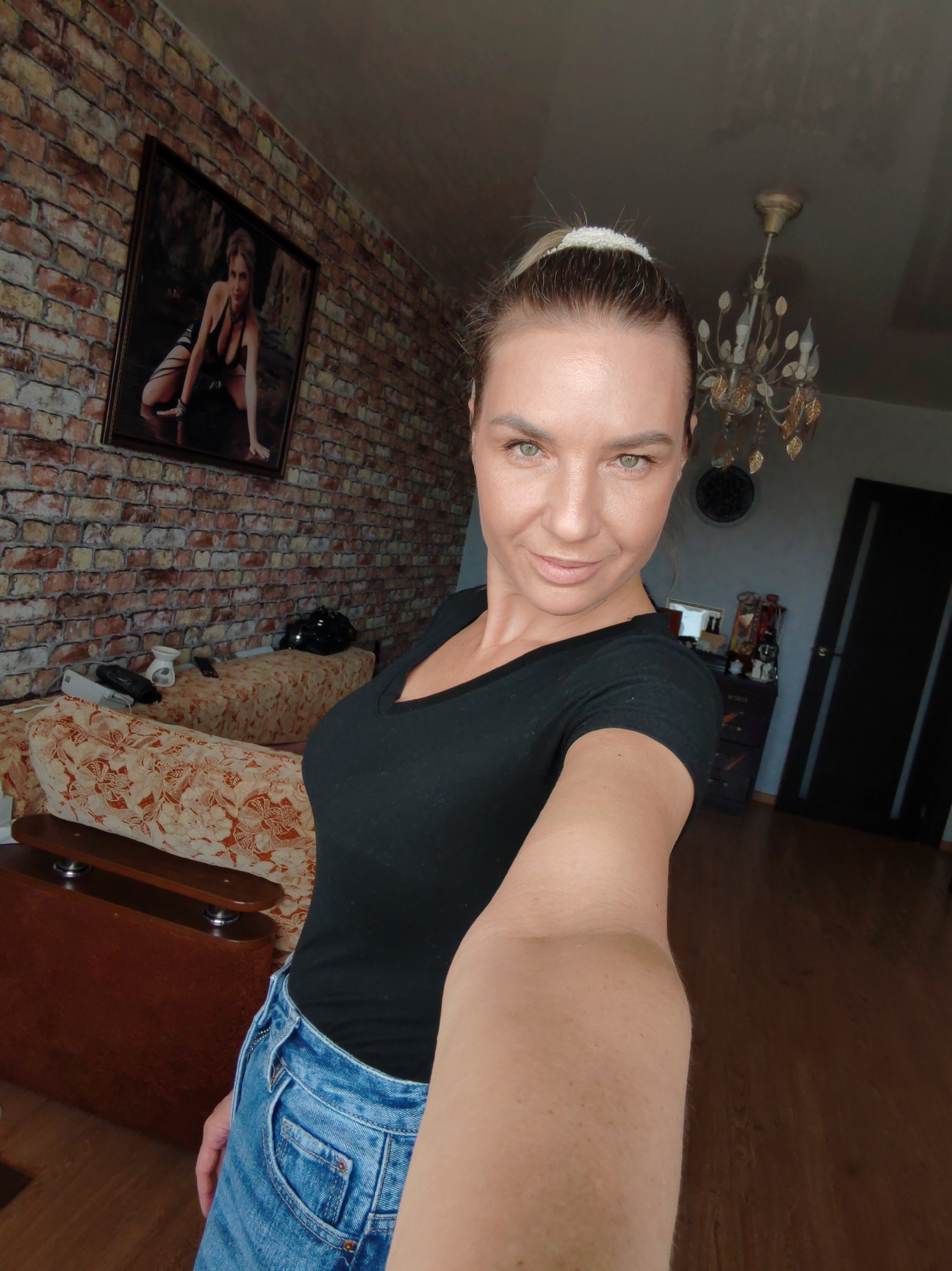 An adult female is depicted taking a selfie inside a residence in Petropavlovsk-Kamchatsky, Russia. The subject, appearing to be in her 30s or 40s, has light skin, green eyes, and light brown hair secured in a high ponytail with a white scrunchie. She wears a black V-neck t-shirt and blue denim jeans, holding the camera with her right arm extended, and displaying a slight smile. The background on the left features a wall covered in red brick-patterned wallpaper. A framed picture depicting a woman in a black halter-top and garters, kneeling on all fours, is mounted on this wall. Below the picture, a sofa upholstered in an orange and brown floral pattern is present. On the sofa, a black motorcycle helmet, a white ceramic essential oil diffuser, and other indistinct objects are visible. The ceiling is light-colored and reflective. A gold-toned, multi-branched chandelier with leaf-shaped embellishments and several light bulbs hangs from the ceiling in the upper right quadrant. To the right, a dark wooden dresser with various items on its surface is partially visible, along with a dark circular wall ornament or clock. A dark brown or black interior door with two vertical panels and a metallic doorknob occupies the far right side of the image. The floor is constructed of wood panels.